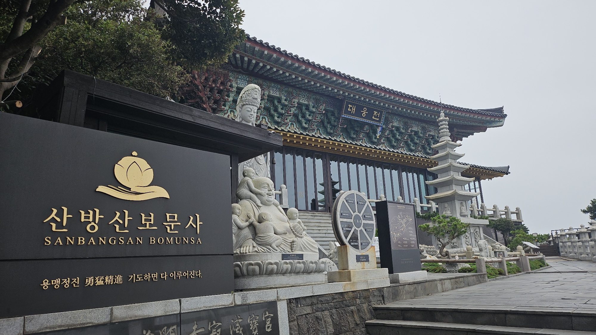 The image captures a view of Sanbangsan Bomunsa temple in Seogwipo-si, South Korea. In the foreground, a large black sign displays the temple's name in Korean and English, "SANBANGSAN BOMUNSA," along with a golden lotus flower logo. Below the name, a Korean inscription reads, "용맹정진 勇猛精進 기도하면 다 이루어진다." To the right of the sign, a stone statue of a laughing Buddha surrounded by children is visible. Further back, a large, ornate temple building with traditional Korean architecture stands, featuring colorful decorative carvings and a multi-tiered pagoda. Stone railings and pathways lead towards the temple. The sky is overcast, and the overall atmosphere is serene.