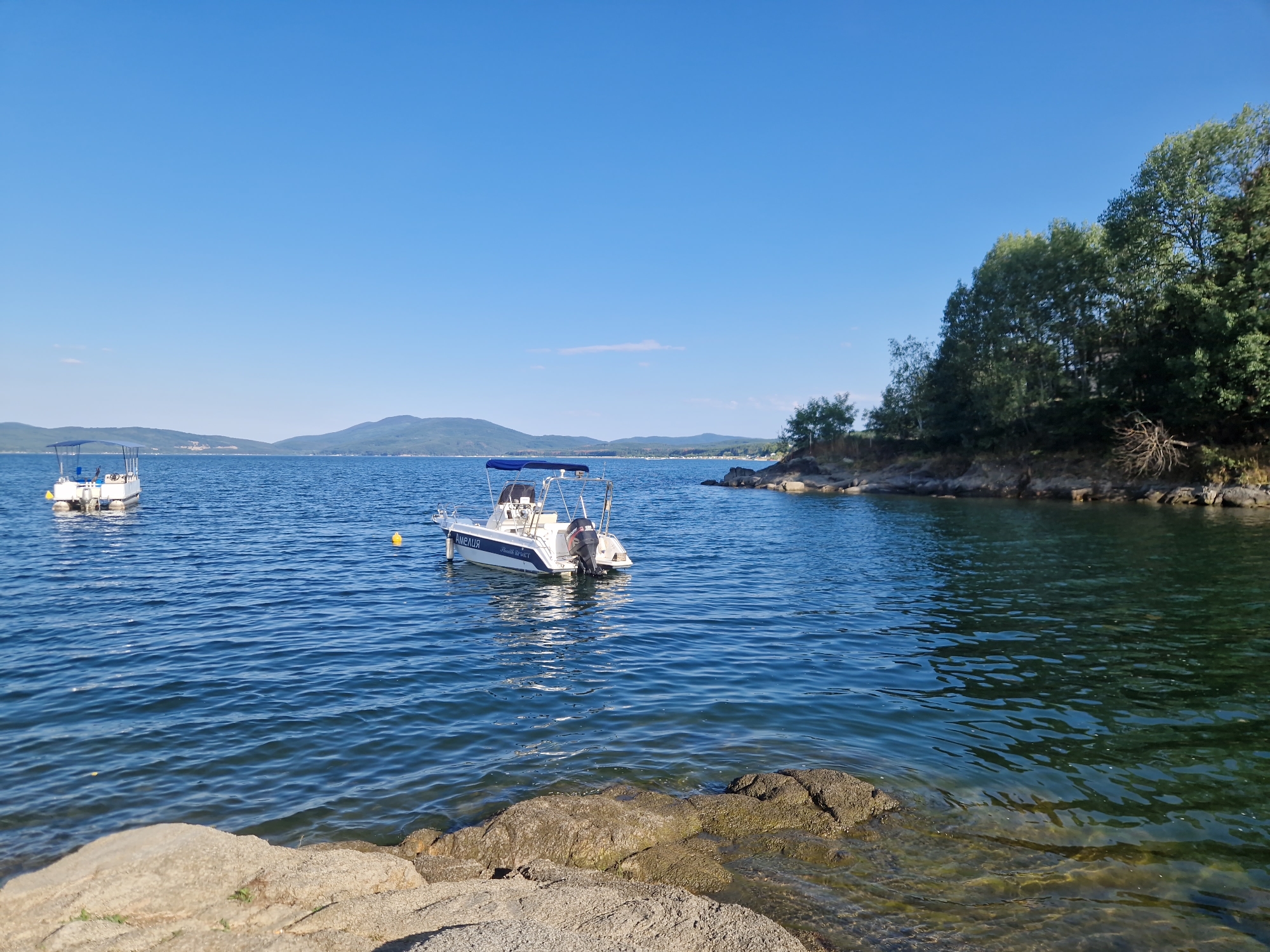 The image depicts a serene lake scene in Dolni Pasarel, Bulgaria, under a clear blue sky. In the foreground, rocky shorelines are visible, with some rocks submerged in the clear, rippling water. Two white motorboats are anchored in the lake. One boat, closer to the right side of the image, has the name "AMEЛIЯ" printed on its hull. The second boat, further to the left, appears similar in design. The background features rolling hills covered in lush green trees, characteristic of the landscape surrounding Dolni Pasarel. The overall atmosphere is tranquil and idyllic.
