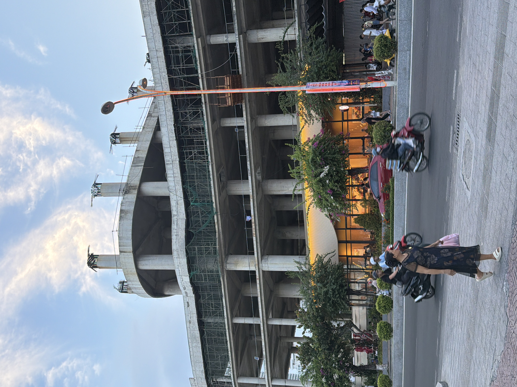 The image depicts a street scene in Nha Trang Ward, Vietnam, dominated by a large, unfinished concrete building. The building appears to be under construction, with exposed rebar and scaffolding visible on its upper levels. On the ground floor, a section of the building has a glass facade with warm interior lighting, suggesting it may be a restaurant or commercial space.
In the foreground, a woman in a dark floral dress walks on a paved sidewalk, carrying a light pink bag. Several motorcycles are in motion on the street, including one with a driver and a passenger, and another with a single rider. A red car is parked on the side of the road.
A distinctive street lamp with multiple lights stands tall near the edge of the sidewalk. Trees with green foliage and some flowering plants line the street. The sky is a blend of blue with scattered white clouds, and the lighting suggests it is either early morning or late afternoon. The overall scene captures a moment of urban activity in a developing area.