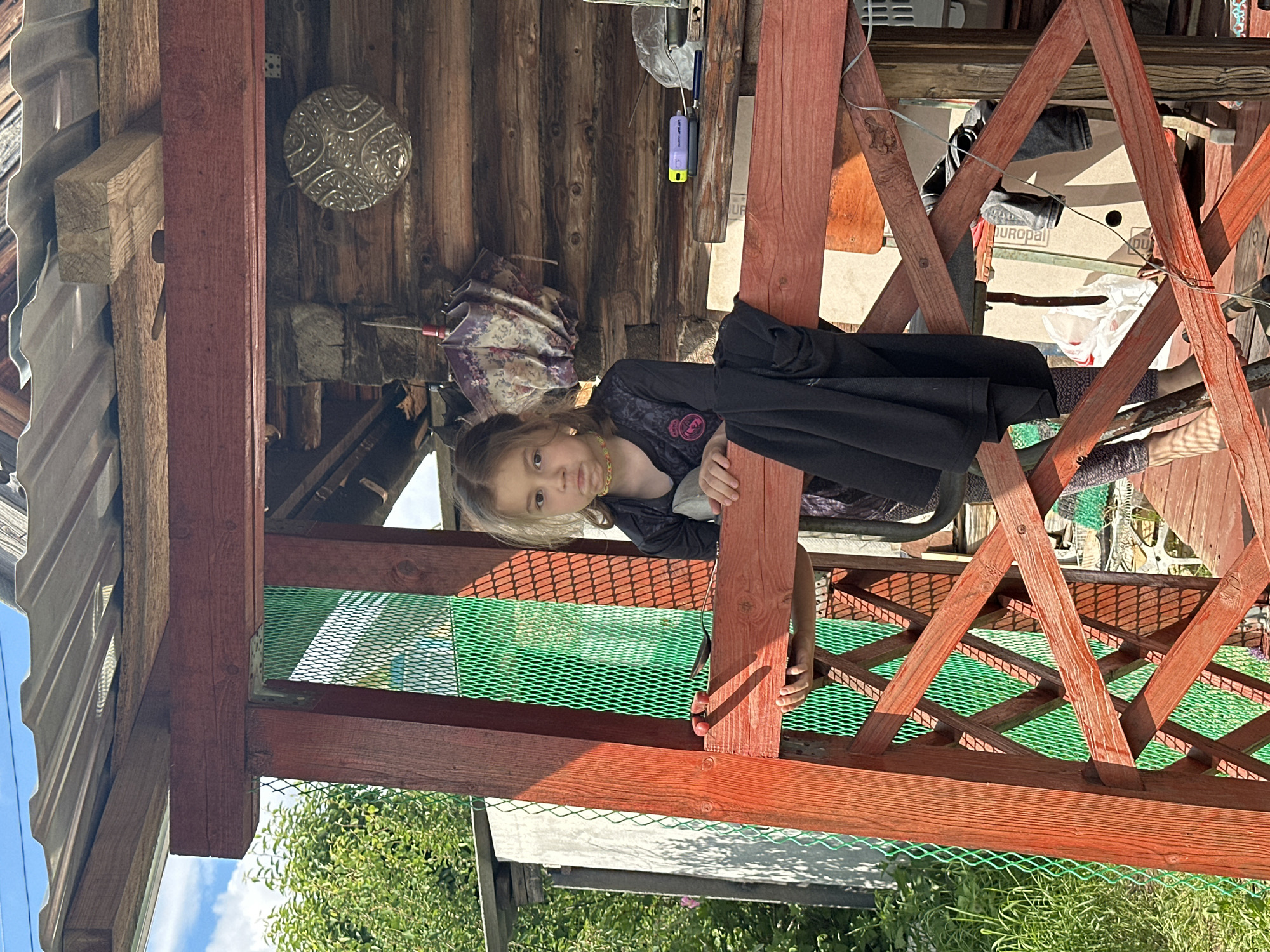 A young girl stands inside a wooden structure in Khokhryaki, Russia. The structure is painted red and has green netting along the bottom. The background includes greenery and a wooden building with various objects hanging on the wall. The girl is wearing a dark outfit and has light brown hair. She is looking at the camera.