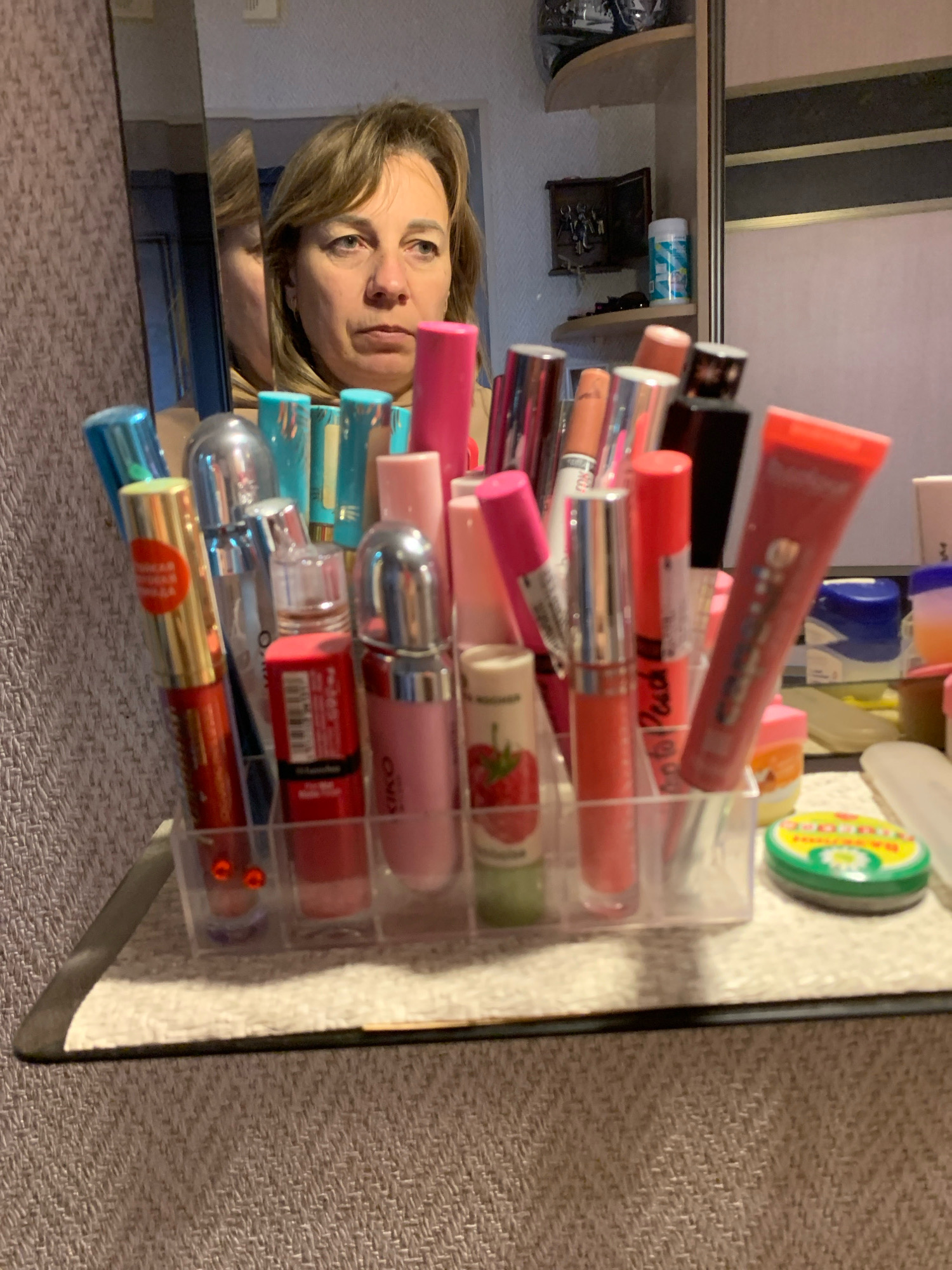 In this close-up, medium-angle shot, a woman is reflected in a mirror, her gaze directed slightly downward. She has shoulder-length blonde hair and is looking straight ahead with a neutral expression. In the foreground, a clear plastic organizer holds a collection of lip glosses and lipsticks. The products are varied in color, with shades of pink, red, and orange being prominent. Some of the lip gloss tubes are clear, revealing the product inside, while others are opaque. The organizer is placed on a textured, light-colored surface, which could be a mat or fabric. Behind the organizer and to the right, other cosmetic items and small containers are visible on a shelf. The background consists of a wall with a patterned texture and a sliver of a wardrobe or cabinet. The overall lighting is artificial and somewhat dim. There is no indication that this scene is located in Minsk, Belarus.
