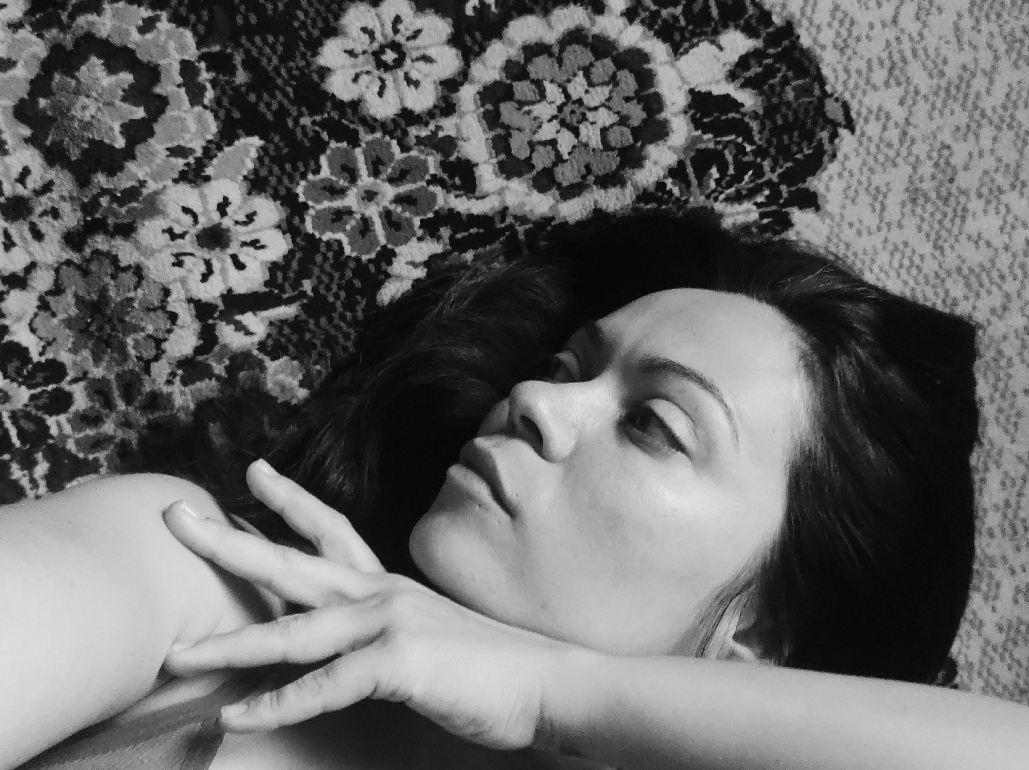A black and white close-up photograph shows a woman lying down with her head turned to the left. Her eyes are partially closed, and her right hand is resting on her shoulder. She has dark hair. The background consists of a patterned textile. The location is Unknown, Unknown.