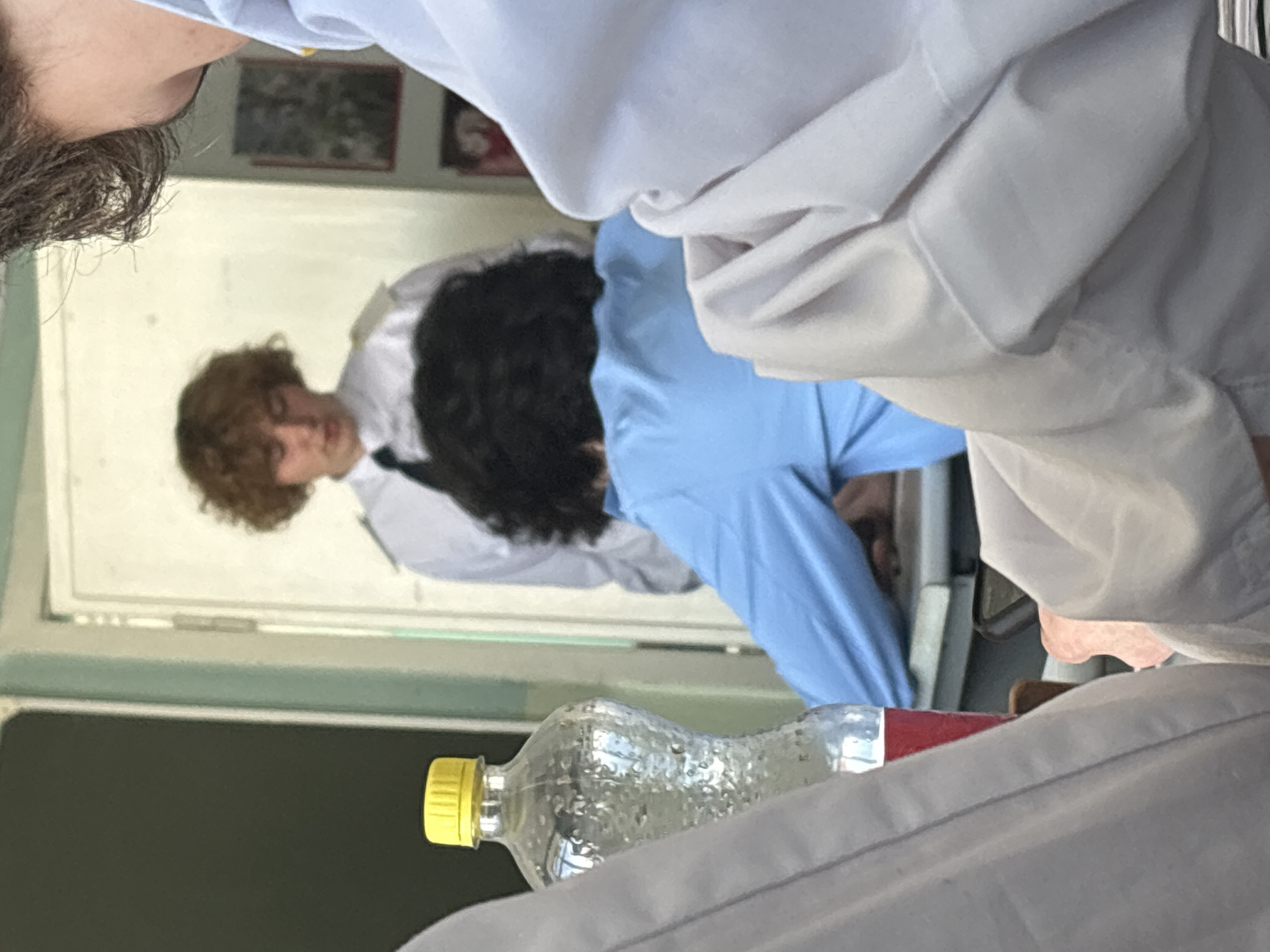 The image is presented on its side, rotated 90 degrees counter-clockwise. It captures an indoor scene featuring multiple individuals and a water bottle.
In the upper left quadrant, partially visible, is the side profile of a person's head and shoulder, with dark, curly hair and wearing a light-colored garment.
Dominating the center of the frame are two individuals. One person, positioned to the right of the other, has light brown, curly hair and is wearing a white collared shirt with a dark tie or vest. They appear to be looking forward or slightly downwards. The second person, mostly seen from behind, has dark, curly hair and is wearing a light blue long-sleeved shirt. This individual is leaning forward, suggesting engagement with an object or activity outside the direct view of the camera.
In the lower right corner, a clear plastic water bottle with a yellow cap and a red label is visible, containing water droplets. It rests on a light-colored surface, possibly clothing or a table.
The background consists of a light-colored wall with two framed pictures hanging on it. The details within the frames are indistinct due to blurriness. The overall lighting is bright and even, characteristic of an indoor setting during the daytime. No discernable text is visible on any objects or in the background. The scene suggests a candid moment, possibly in a classroom or a casual meeting environment.