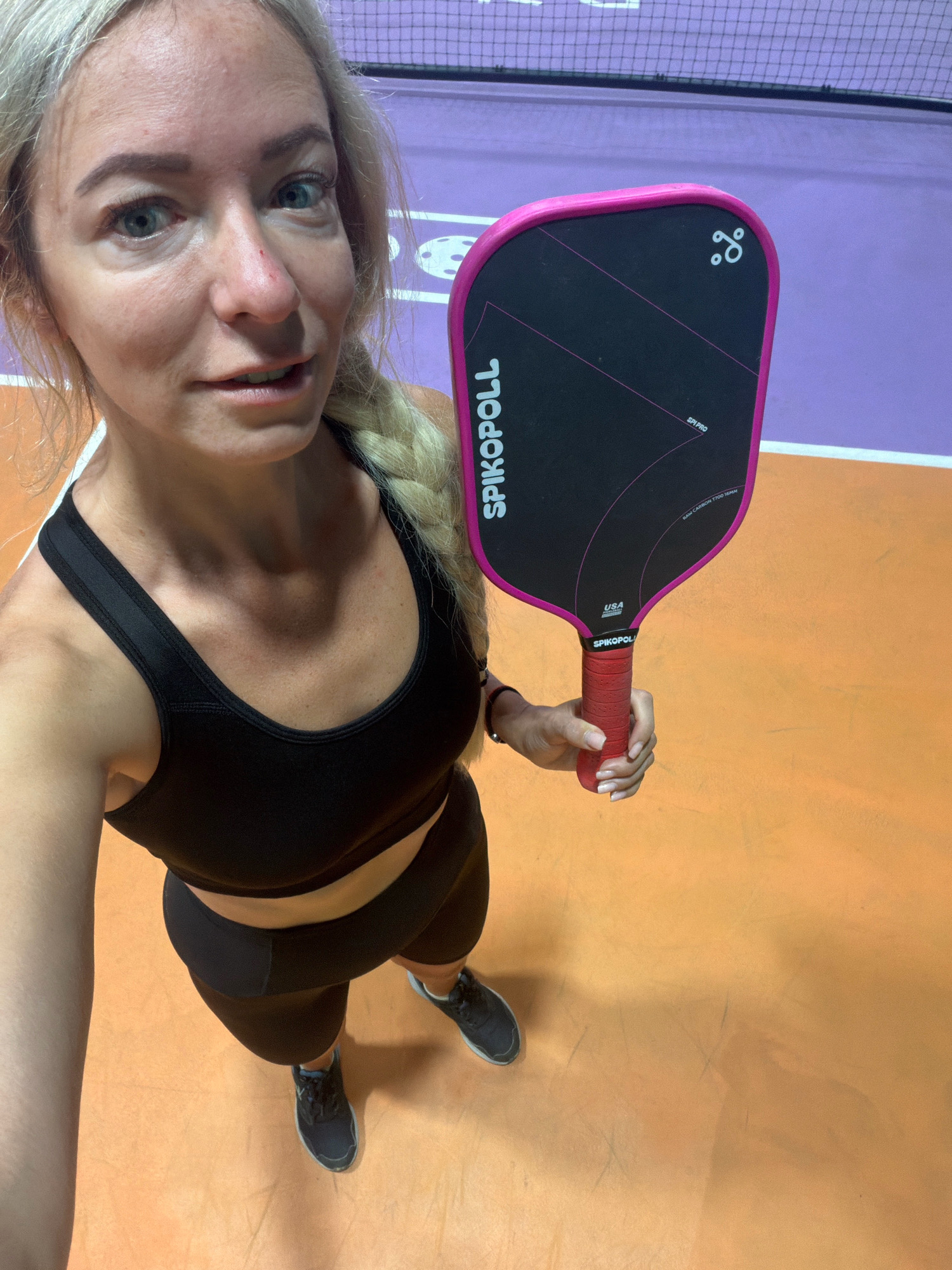 A woman stands on a pickleball court holding a pink and black pickleball paddle. She is wearing a black sports bra and black shorts, with black athletic shoes. The court is marked with orange and purple lines, and a net is visible in the background. The lighting suggests an indoor setting. There are no identifiable landmarks or elements to suggest Da Lat, Vietnam; the scene is generic to an indoor sports facility.