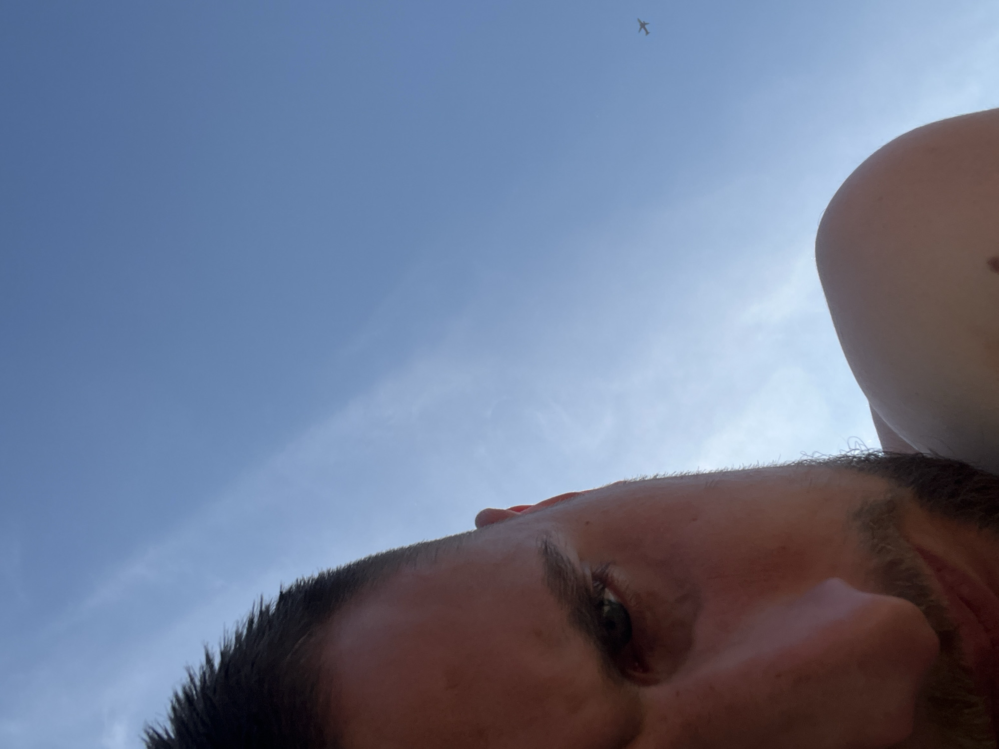 A low-angle outdoor shot depicts the upper-left portion of a male's head and right shoulder in the foreground, set against a clear blue sky in Kapustino, Russia. The man, with short dark hair and a beard, has his right eye prominently visible and appears to be looking towards the viewer. The lower half of his face, including his nose and mouth, is also partially visible. His skin is illuminated by sunlight, casting some shadow. In the upper-right quadrant of the frame, a small airplane is visible in the distance, flying across the bright blue sky, which contains faint, streaky white clouds.