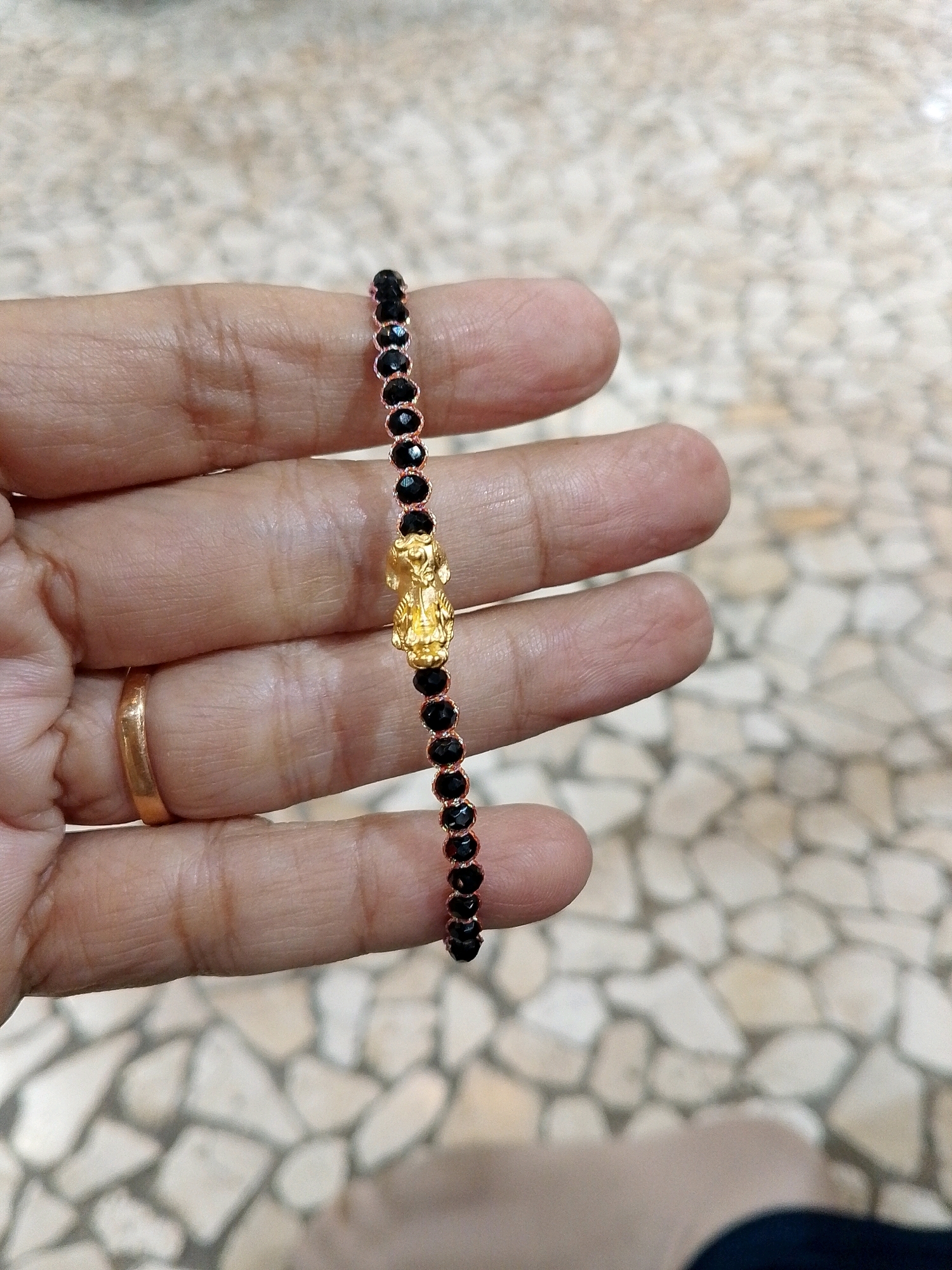 A close-up shot shows a person's hand holding a bracelet. The bracelet features a thin chain with small, black, faceted beads and a golden charm in the center. The charm appears to be of a mythical creature or animal, possibly a lion or a dragon, with intricate detailing. A plain gold band is visible on one of the fingers. The background is blurred, showing a light-colored, textured surface that resembles paving stones. The lighting is soft and natural, suggesting an outdoor or well-lit indoor environment. The image is taken from a slightly high angle, focusing on the hand and the bracelet. The specific location within the city of Makassar, Indonesia, cannot be determined from the provided image.