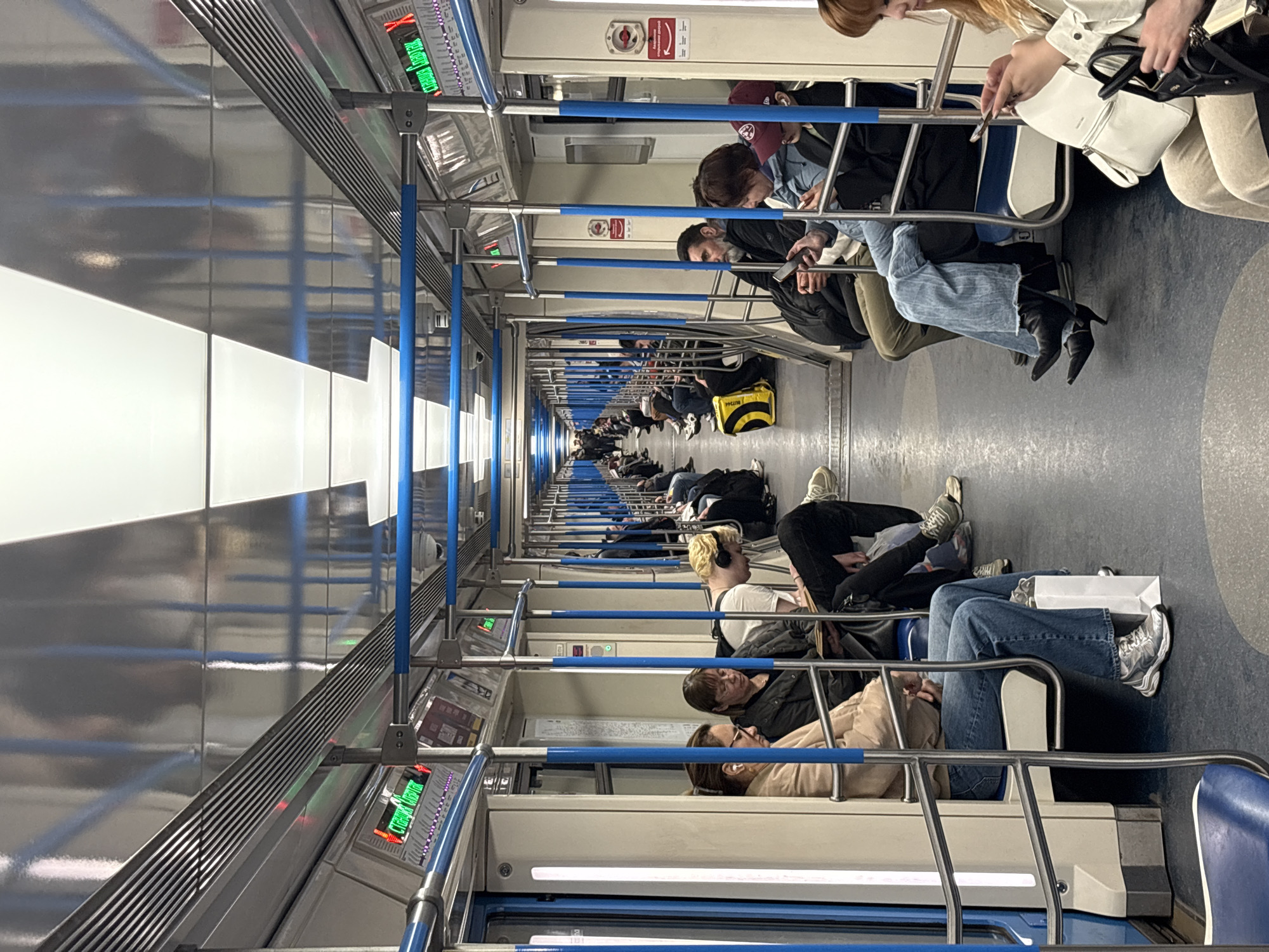 The image shows the interior of a subway car, likely in Khimki, Russia. The car is occupied by numerous passengers seated along both sides of the aisle. Many are using their phones. There are overhead handrails and electronic displays. The car has a metallic and blue color scheme.