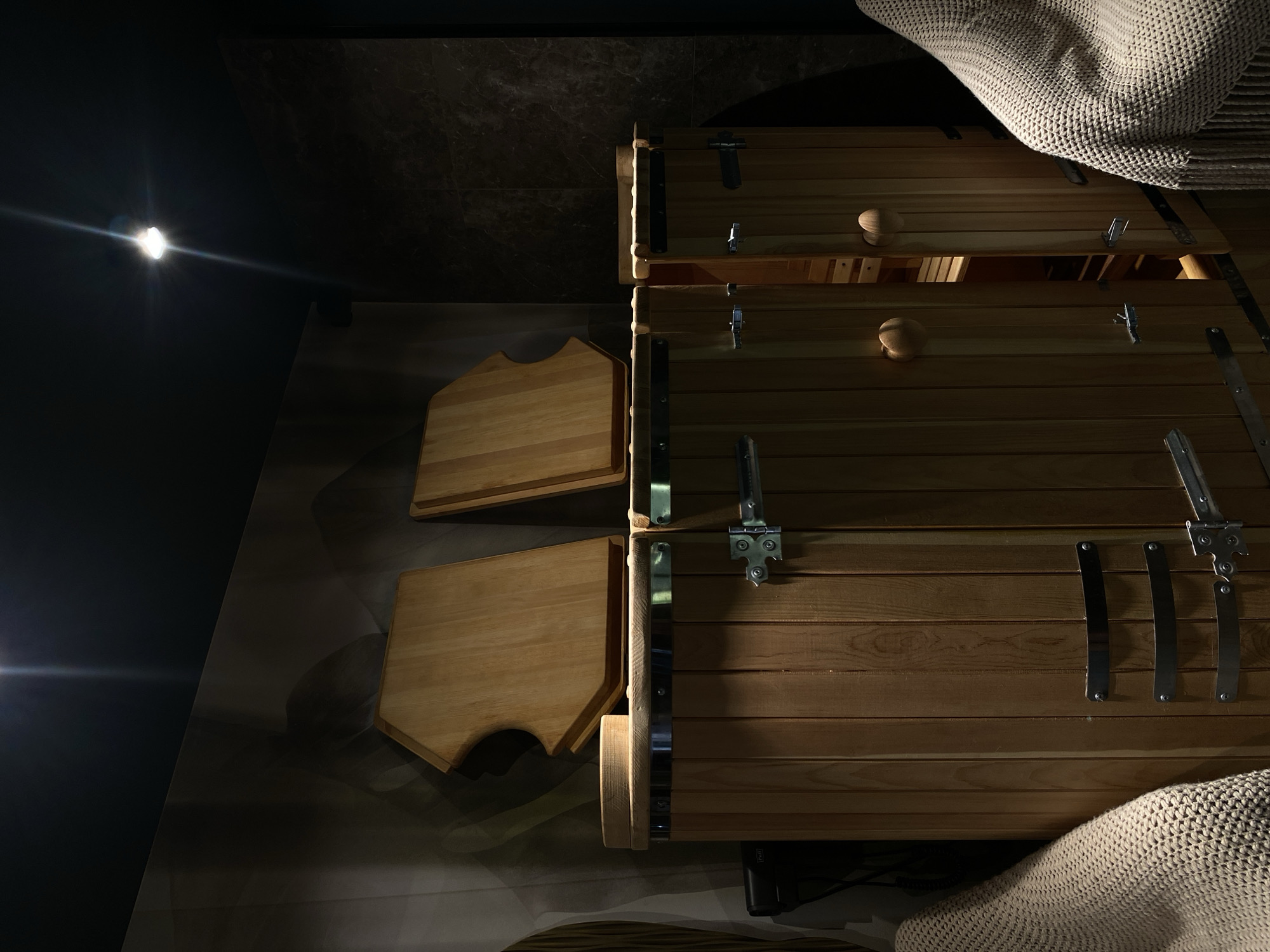 The image shows the interior of a wooden sauna, featuring horizontal planks of wood with metallic hinges and attachments. Two hinged wooden shelves are visible on the left side. A beige, knit blanket is draped over part of the structure on the top right and bottom right. The background transitions from a dark blue wall on the left to a textured, marble-like wall in the center. A light source is visible on the upper left, casting a glare. The location, Votkinsk, Russia, is not deducible from the image.