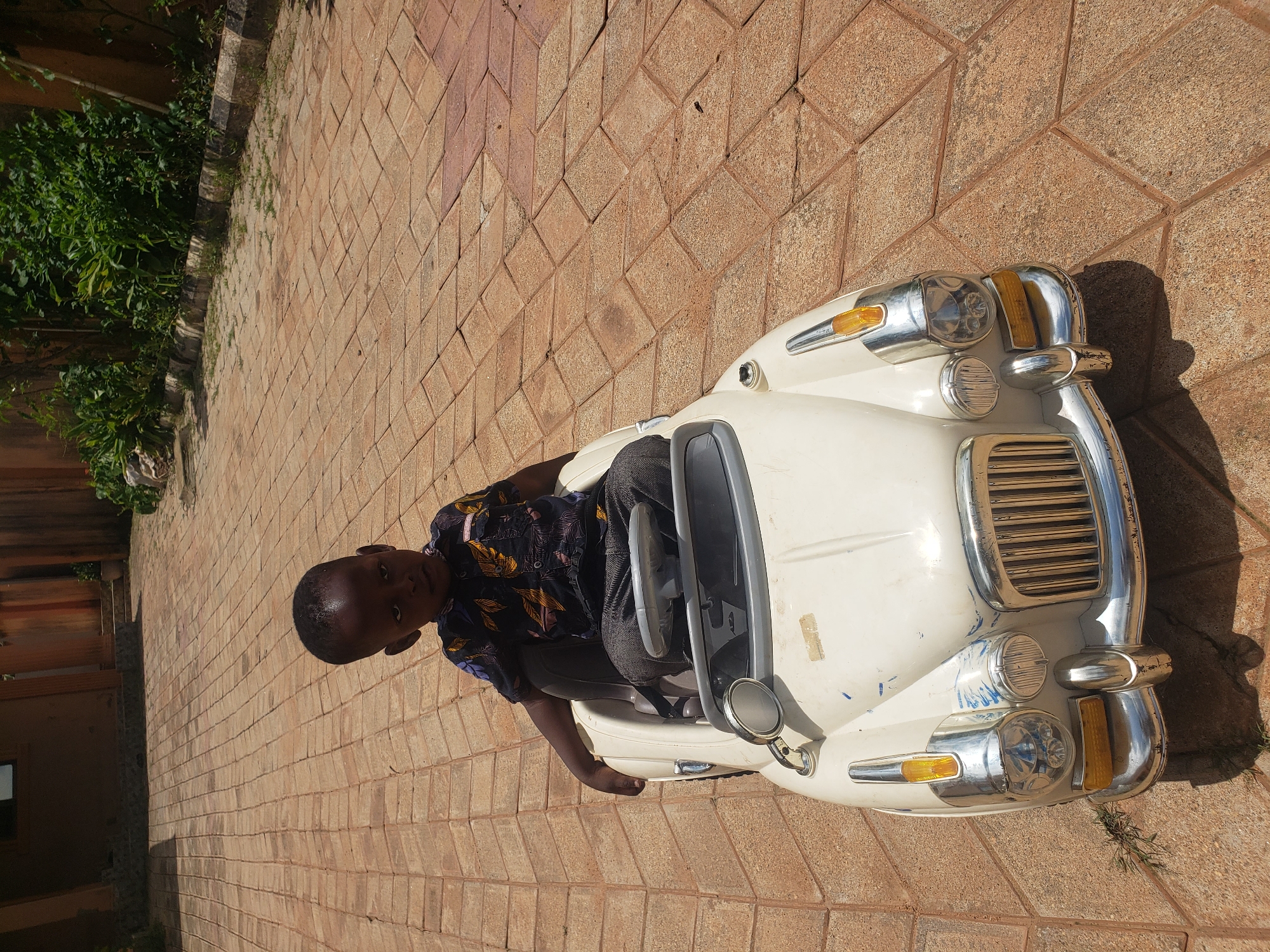 A young Black boy is seated in a white toy car on a paved outdoor area with a reddish-brown tile pattern. The boy is wearing a dark short-sleeved shirt with a yellow and white floral pattern and dark pants. He is looking towards the right side of the frame, with his left hand resting on the edge of the car. The toy car is detailed with a chrome grille, headlights, and a side mirror. The background shows a building with a brown facade and lush green foliage. The scene is set in Sabon Tasha, Nigeria.