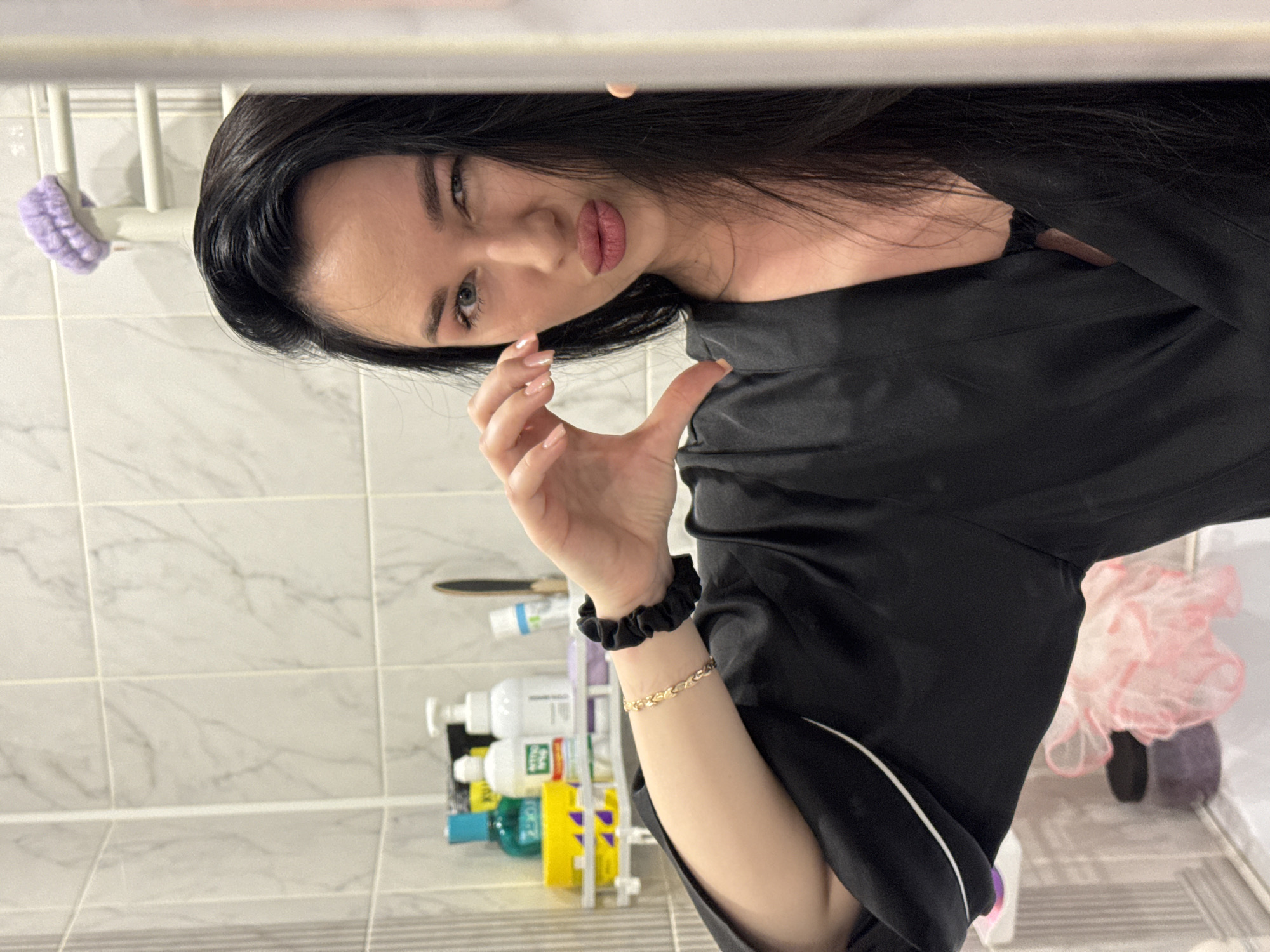 A young woman with long, dark hair is taking a selfie in a bathroom. She is wearing a black satin robe with white trim. She is winking and making a heart shape with her hands. Behind her, there are shelves with various bottles of toiletries, including shampoo and lotion. The bathroom walls are tiled with a marble-like pattern. The overall impression is a casual, personal moment captured in a domestic setting. The location is not specified in the image.