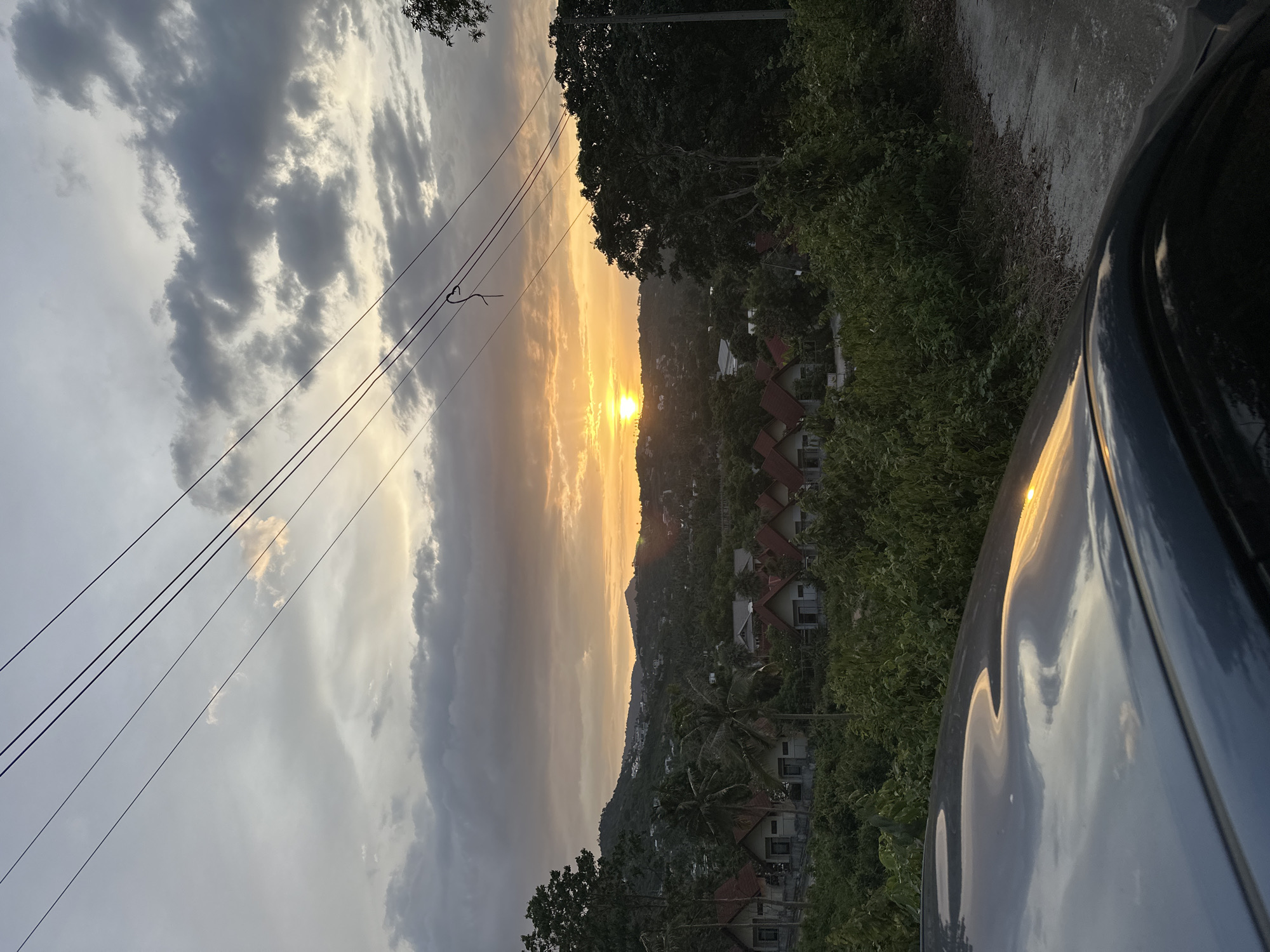 A sunset scene is captured in Ko Samui District, Thailand, featuring a wide view of a tropical landscape. The sky is filled with dramatic clouds, some dark gray and others illuminated by the orange and yellow hues of the setting sun. Multiple power lines are strung across the upper portion of the image. Below the sky, a hilly terrain is covered in lush green vegetation, dotted with houses with red tiled roofs. Palm trees are interspersed among the foliage. In the foreground, the curved, reflective surface of a car's hood mirrors the sky and the surrounding landscape. A dirt or gravel path is visible on the right side of the image. The overall atmosphere is serene and picturesque.