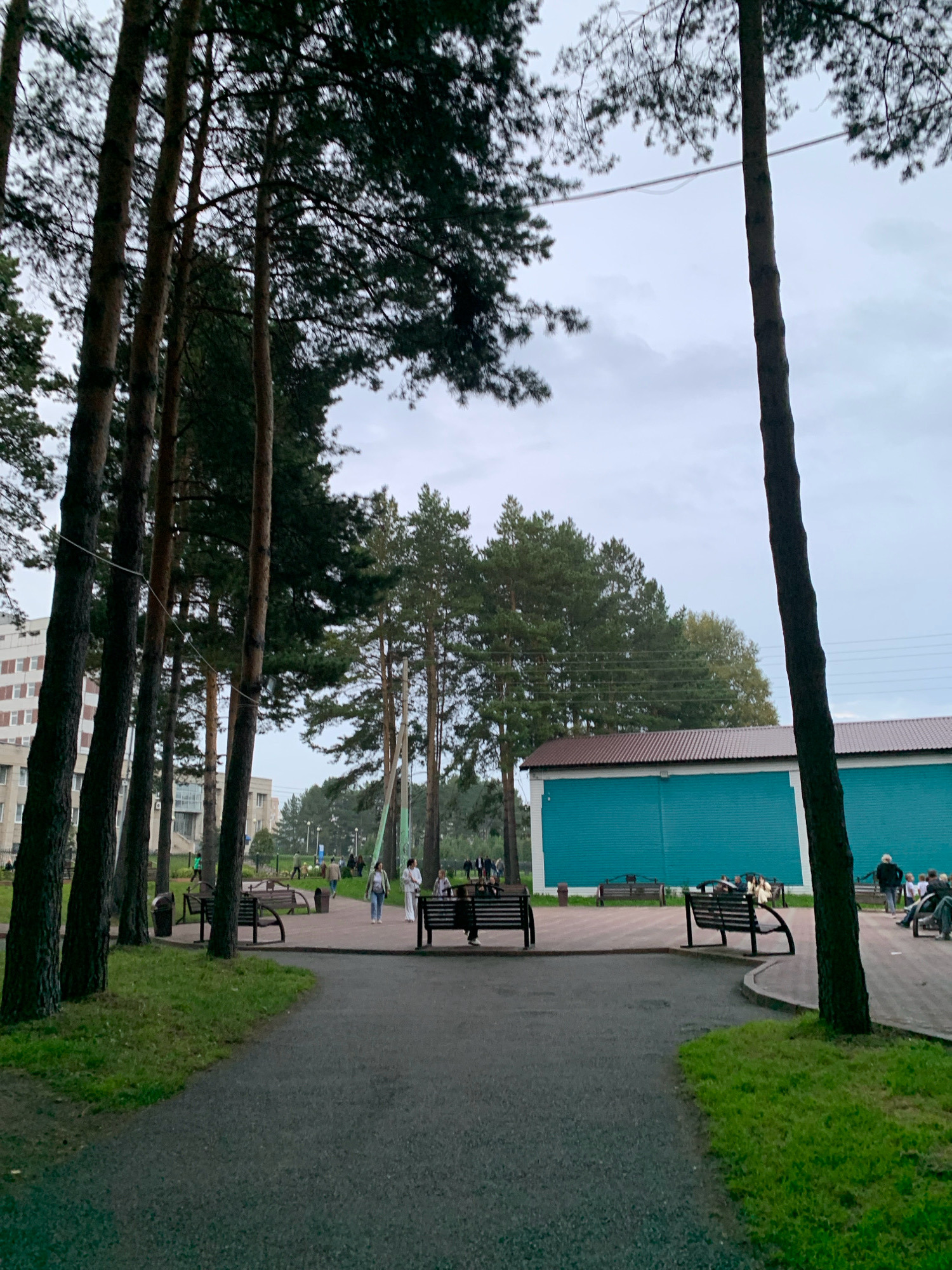 The image depicts an outdoor scene in what is identified as Prokopyevsk, Russia. In the foreground, a paved pathway curves through a grassy area dotted with tall pine trees. Several park benches are visible along the pathway. In the mid-ground, people are walking and sitting on benches. Behind the pathway, there is a long, teal-colored building with a brown roof. In the background, more trees and buildings can be seen, including a multi-story building. The sky is overcast.
