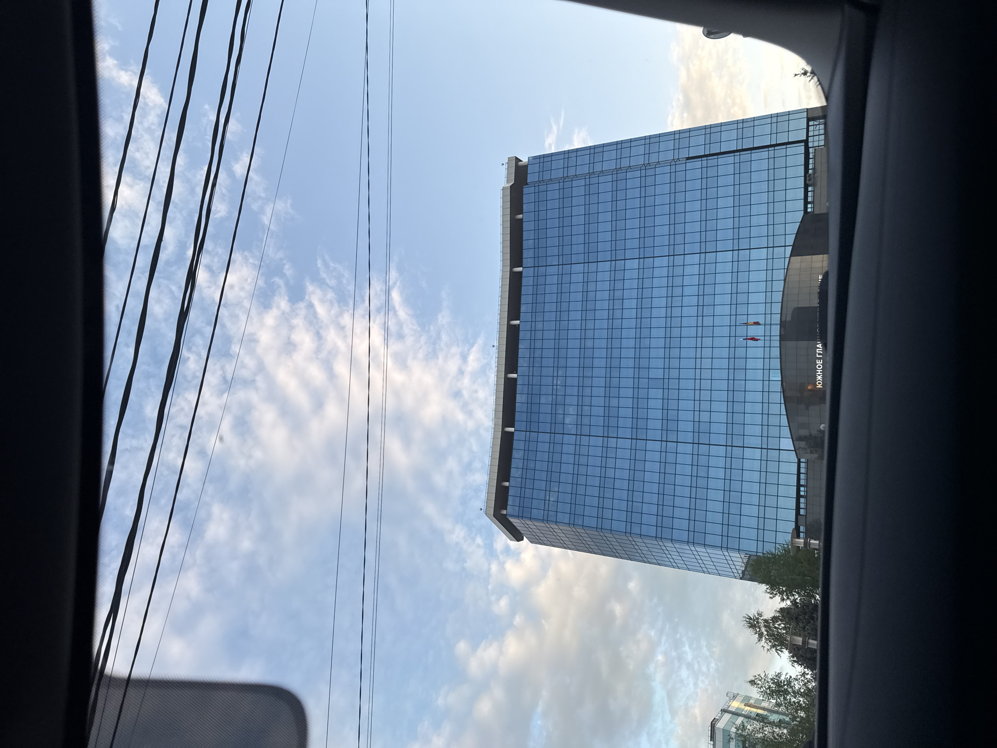 The image shows a tall, glass-fronted building with a grid-like pattern of windows, viewed from inside a vehicle. A clear blue sky with scattered clouds serves as the backdrop, interspersed with power lines on the left. The building has a modern design and the name "ЮЖНОЕ ГЛА" (likely the name of the building or an organization) is visible on the side, along with two small flags. Some greenery and a glimpse of another building are visible near the base of the main building. The shot appears to be taken in a city environment. The location details given are Unknown, Unknown.