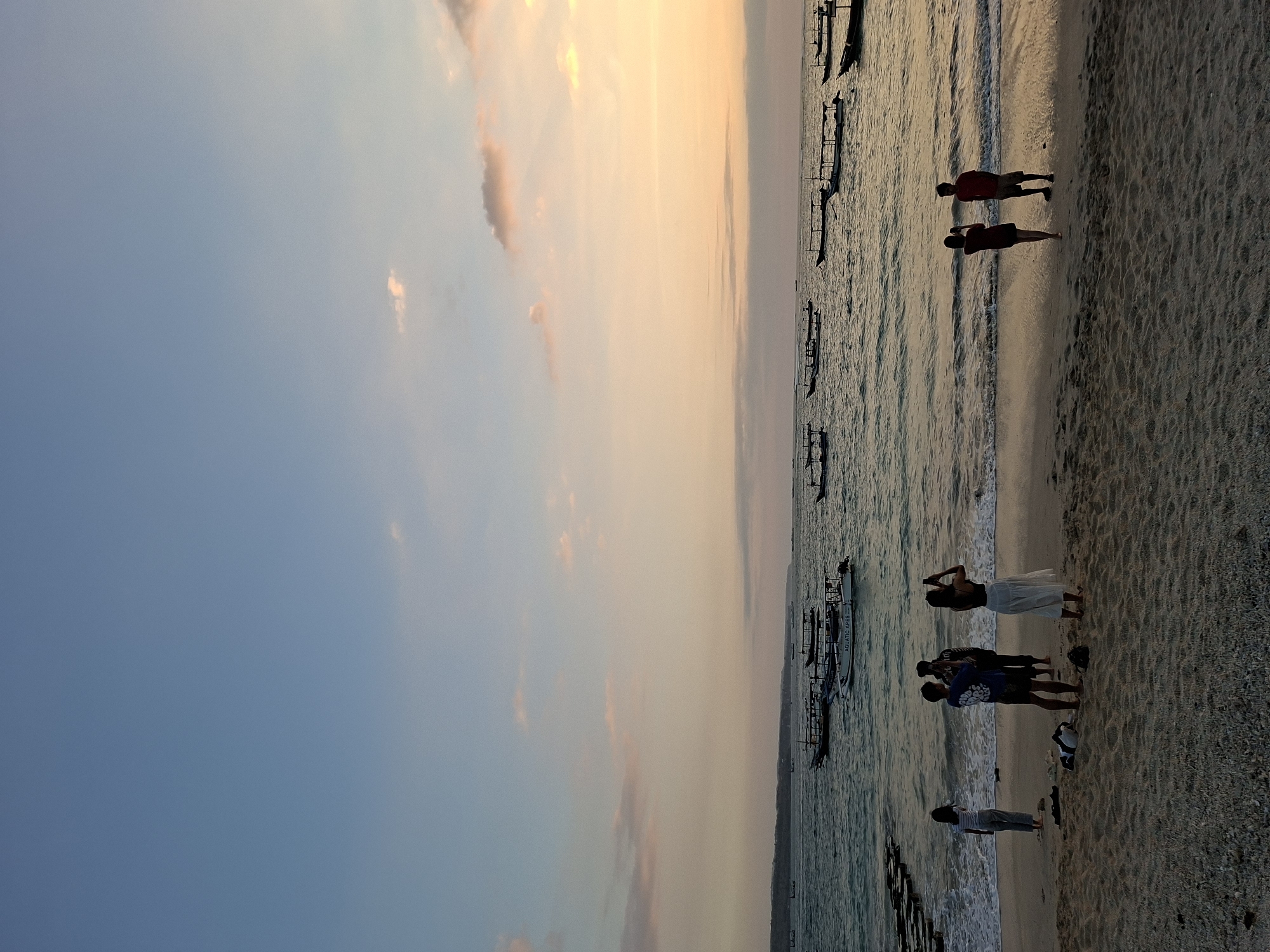 A wide shot captures a beach scene in Kuta, Indonesia, at dusk. The sky is a gradient of soft blues and oranges, with a few wispy clouds. The ocean is calm with gentle waves lapping at the shore. Several traditional Balinese fishing boats, known as jukung, are anchored offshore, their silhouettes visible against the water. On the sandy beach, a group of people are standing, some taking photos with their mobile phones. The sand is a light brown, with small pebbles scattered across its surface. In the foreground, the sand is rippled from the tide. The overall atmosphere is peaceful and serene, evoking a sense of tranquility as the day draws to a close.