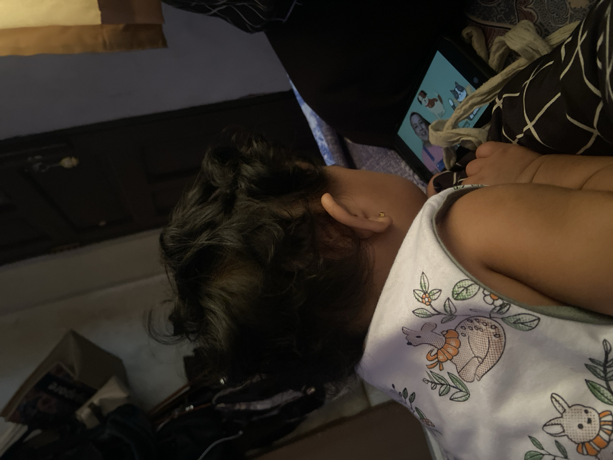 A young child lies on their back, seemingly watching a digital tablet. The tablet screen displays a person and cartoon images. The child wears a white tank top with an animal pattern. The setting is indeterminate and the location in Kuta, Indonesia is not discernible from the image.