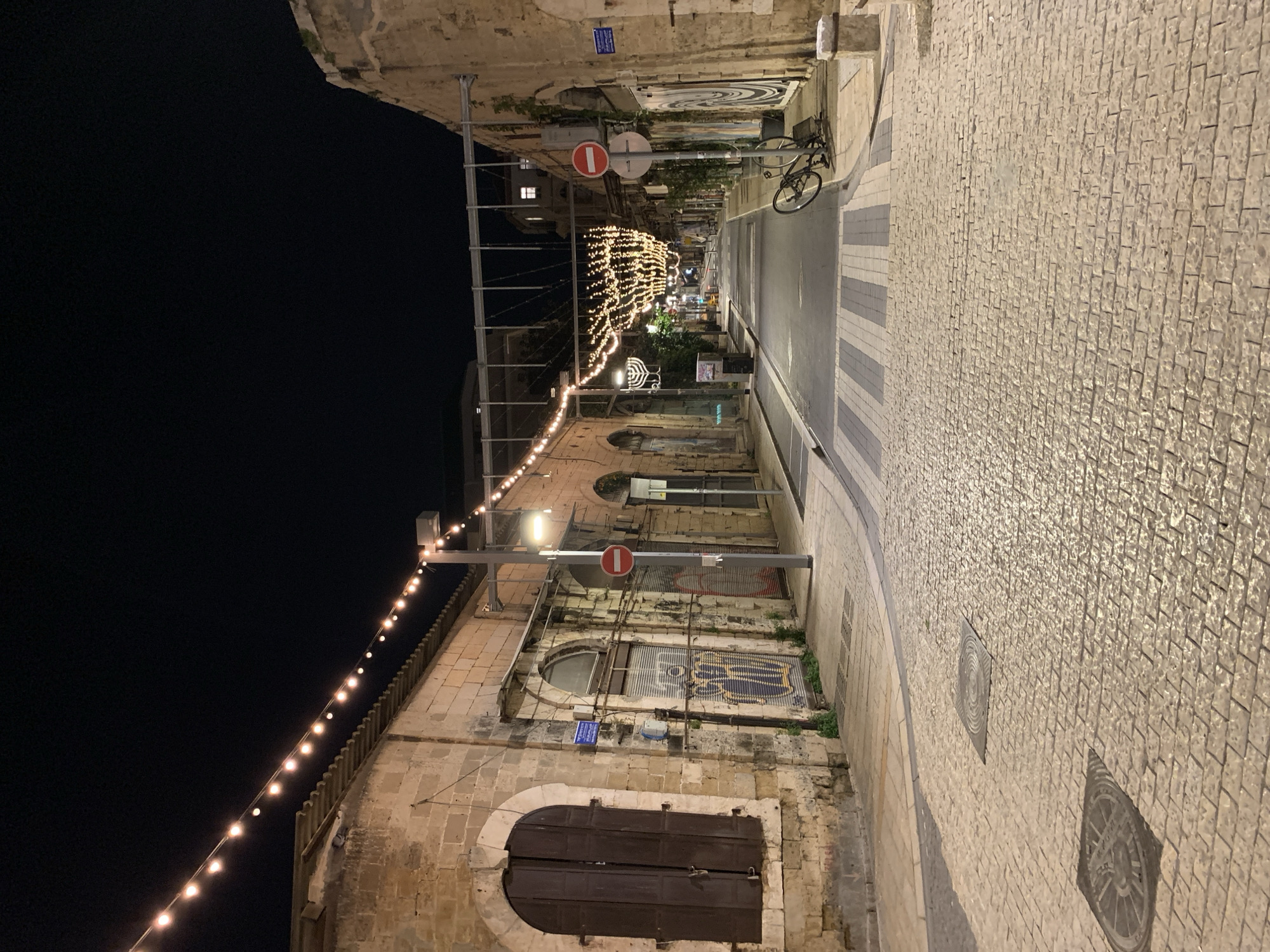 The image displays a deserted cobbled street at night in the Old City of Jaffa, Tel-Aviv, Israel. The street is bordered by two-story buildings constructed from light-colored stone blocks, exhibiting arched window and door openings. Some ground-floor openings are secured by dark wooden shutters or metal roll-down gates, with visible graffiti on several. The scene is illuminated by numerous strings of warm-toned bulb lights suspended overhead and along the building facades. A large, illuminated decorative Hanukkah menorah stands on the left side of the street in the midground.
The street surface comprises light-colored cobblestones, transitioning into a darker asphalt section with a marked crosswalk further down the street. A black bicycle lies on its side on the right-hand sidewalk, adjacent to a metal bicycle rack. Two red-and-white "Do Not Enter" signs are mounted on poles, one on each side of the street near the foreground. Additional streetlights contribute to the illumination. No individuals, actions, or interactions are present in the frame.