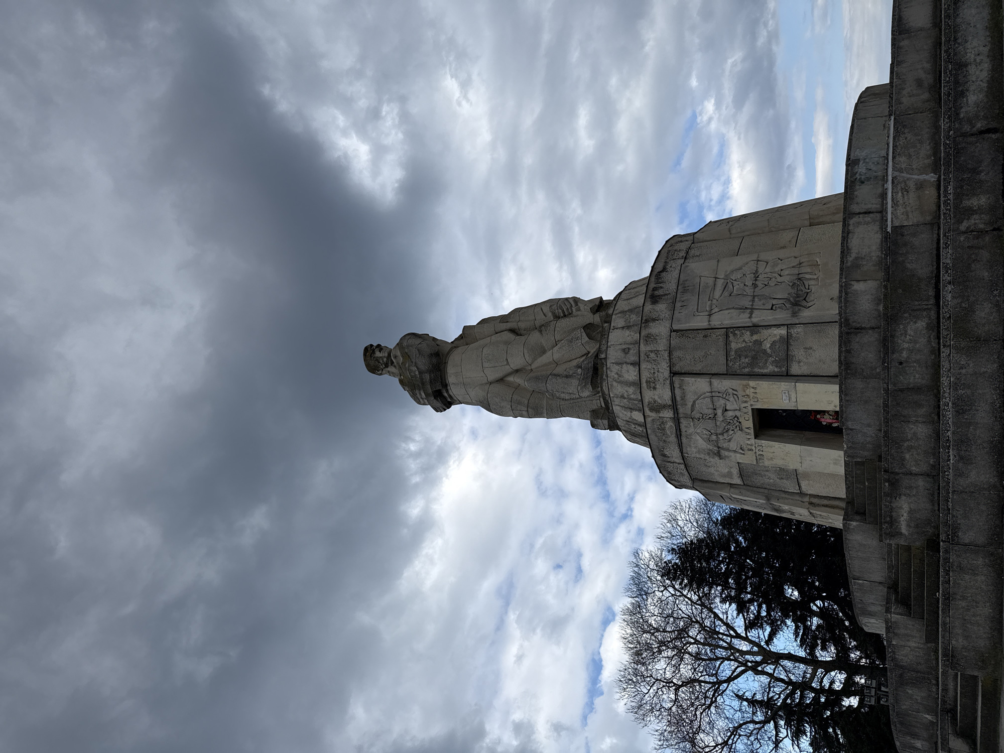 The image depicts the Monument of the Soviet Army in Varna, Bulgaria, taken from a low angle looking upwards. The sky is filled with a mix of dark, ominous clouds and patches of blue sky. The statue of a soldier stands atop a multi-tiered, cylindrical stone monument. The monument features reliefs and inscriptions in Cyrillic. Below the monument, a partially visible tree with bare branches and a dark green conifer are present. Part of the structure includes steps leading up to what appears to be a doorway or entrance. A person is standing inside the doorway.