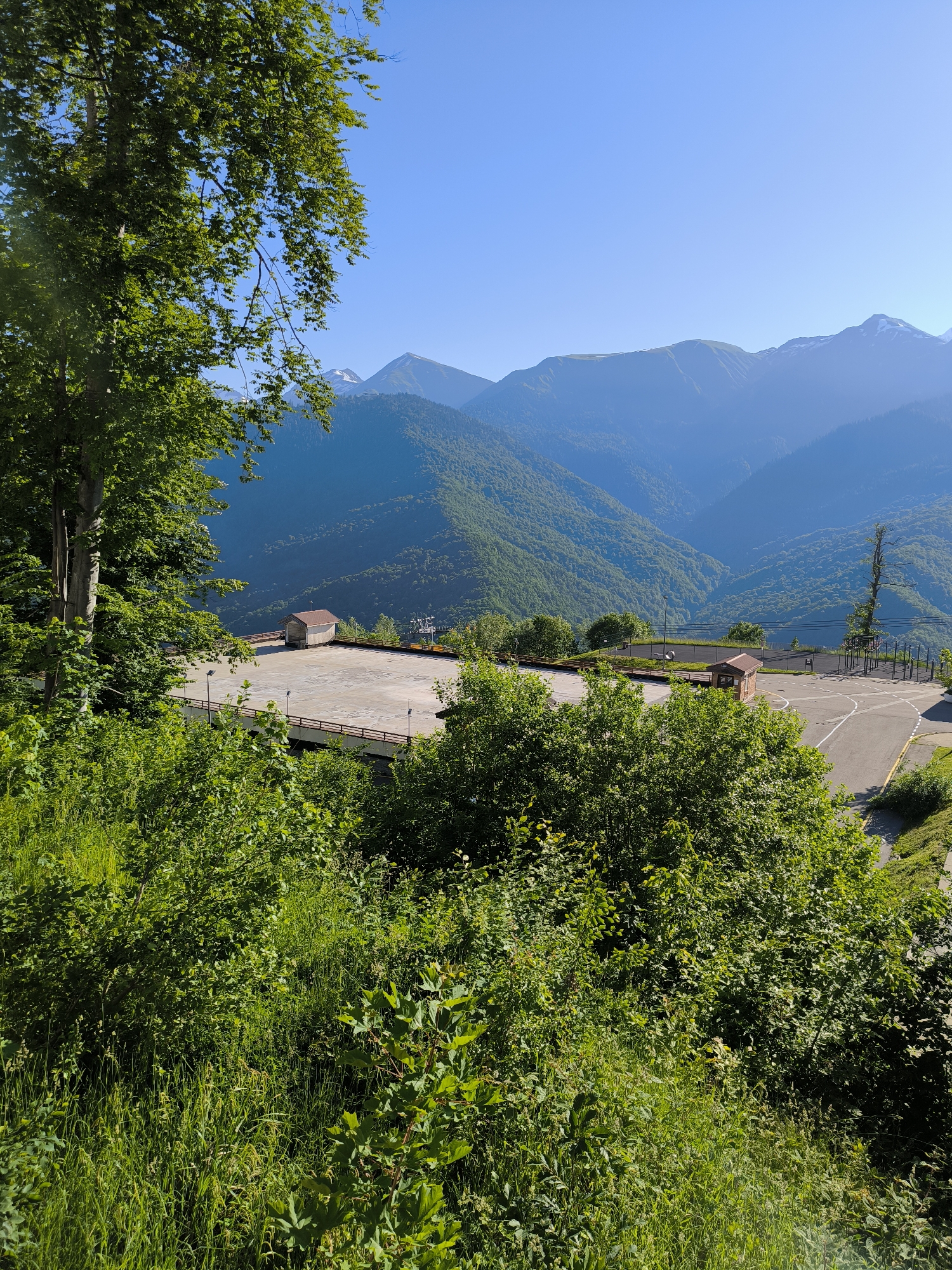 The image captures a vast mountain landscape under a clear blue sky. In the foreground, lush green trees and vegetation dominate the lower portion of the frame, with a large tree on the left providing partial obstruction of the view.
Beyond the foliage, a paved area, possibly a sports court or a viewing platform, is visible. It features a small, brown wooden structure and a few lamp posts. A railing runs along the edge of this elevated area. In the distance, rolling green mountains stretch out, their slopes covered in dense forest. Further still, majestic, snow-capped peaks rise against the horizon, indicating a significant altitude. The overall scene suggests a natural park or resort area in a mountainous region.
This scene is likely located in Estosadok, Russia, known for its stunning mountain scenery and recreational facilities, including those at the Krasnaya Polyana ski resort. The presence of a paved, cleared area with structures suggests a developed tourist or sporting complex amidst the natural beauty of the Caucasus Mountains.