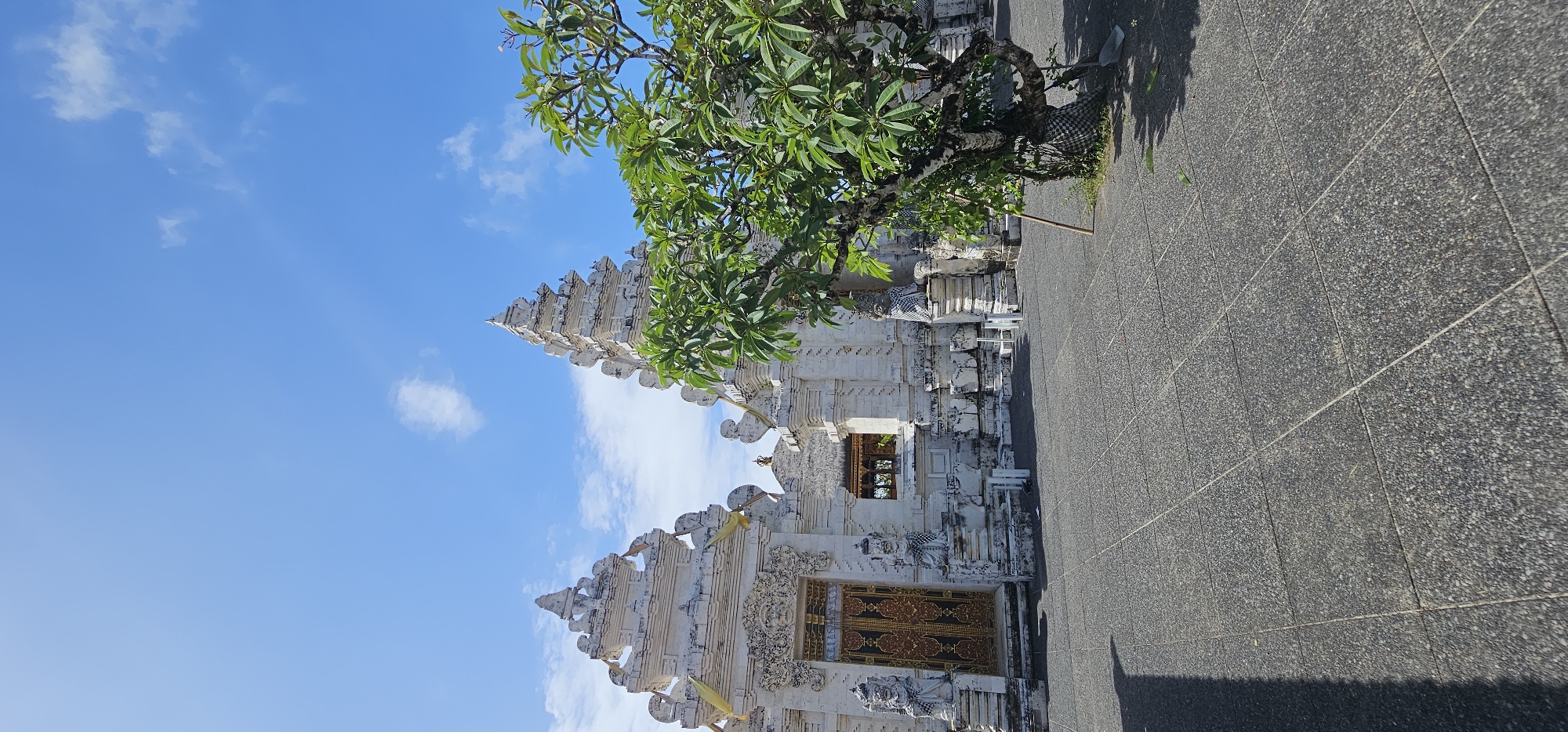 The image depicts a Balinese temple structure in Kuta Selatan, Indonesia. The temple is constructed of white stone with intricate carvings and tiered roofs. A tree with green foliage grows beside the temple, partially obscuring the structure. The sky is blue with scattered white clouds. A paved grey walkway extends from the temple towards the foreground. No people are visible in the frame.