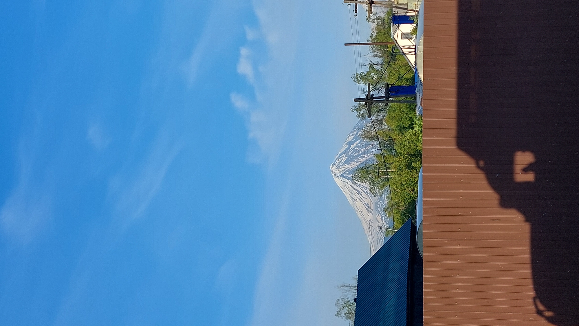 The image depicts a scenic view with a snow-capped mountain in the background, likely Mount Koryaksky, a stratovolcano located in Kamchatka Krai, Russia, near the city of Koryaki. The sky is a clear blue with scattered white clouds. In the foreground, a brown wooden fence runs horizontally across the bottom of the frame, casting a shadow that appears to be from a vehicle or structure. To the left of the fence, a portion of a dark blue roof is visible, suggesting a building. Beyond the fence and to the right, lush green trees form a natural barrier, with glimpses of some houses and utility poles interspersed. The overall scene is natural and serene, with a prominent natural landmark dominating the distant horizon.