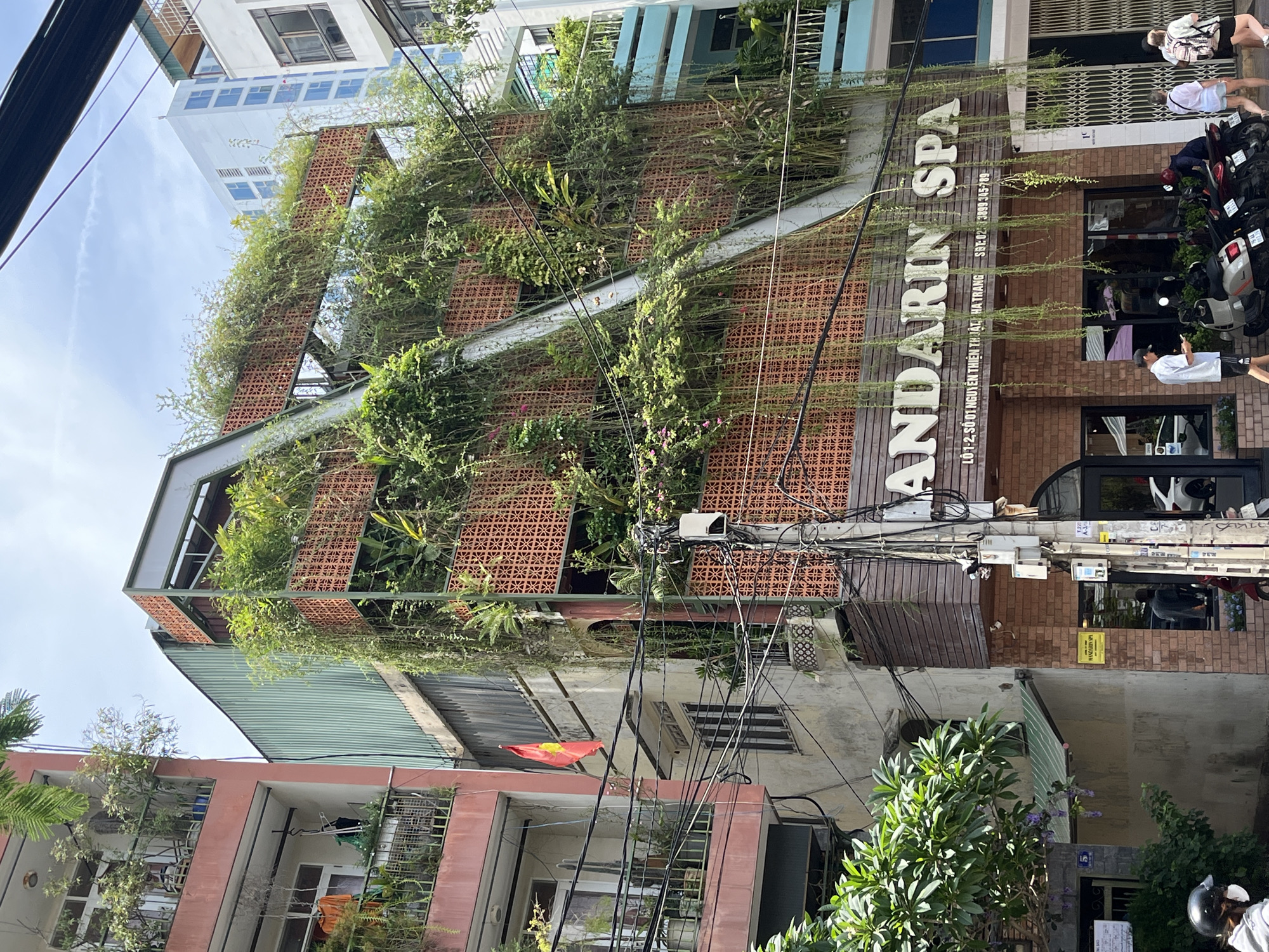 The image shows a multi-story building in Nha Trang Ward, Vietnam, characterized by its prominent green facade covered in dense vegetation. The building features several balconies with plants and hanging vines, and decorative brickwork on its upper levels. A white pipe or structure runs diagonally down the front of the building, integrated with the greenery. Below the vegetated sections, the building has a brick facade with a sign that reads "MANDARIN SPA". Several people are visible on the sidewalk in front of the spa, including a man in a white shirt looking at his phone, and two women with backpacks. Motorcycles are parked along the sidewalk. Other buildings, including a taller, modern white building and a pink residential building, are visible to the sides. Power lines are strung across the scene.