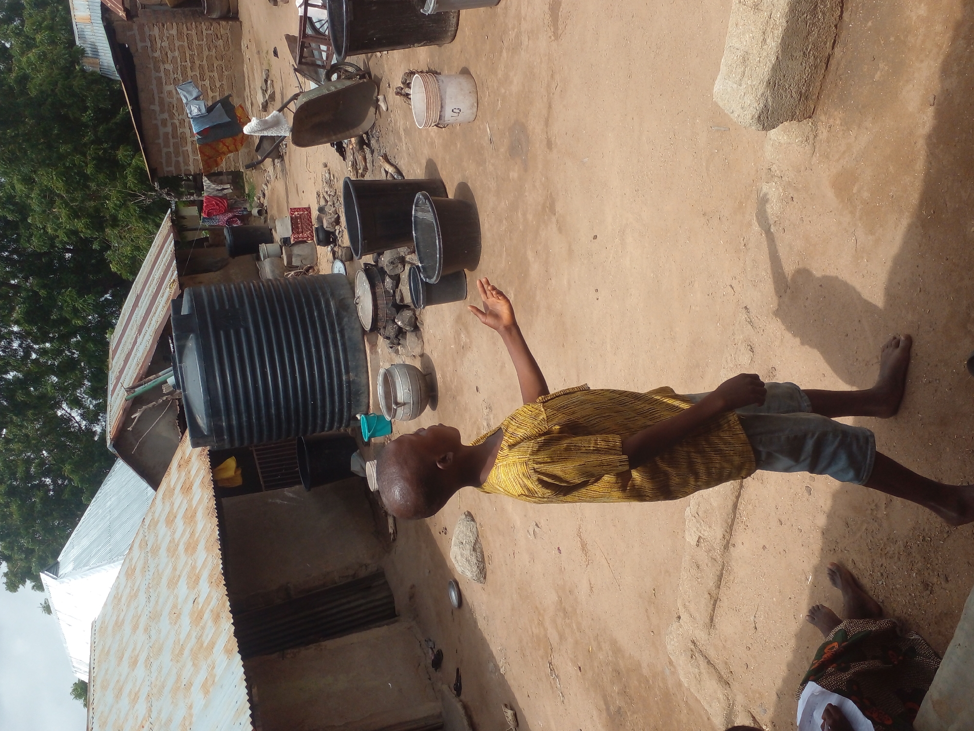 A medium shot captures a young boy with a shaved head, wearing a yellow patterned shirt and blue shorts, standing in a dusty, outdoor village setting. His right arm is extended, palm up, as if reaching for something. He is barefoot. In the background, several rustic structures with corrugated metal roofs and brick walls are visible. A large black water tank stands prominently near the center, and various buckets and containers are scattered around the dirt ground. Clothes are hanging on a line in the middle ground, and a wheelbarrow is visible to the right. The environment appears dry and sunlit, with trees in the background.