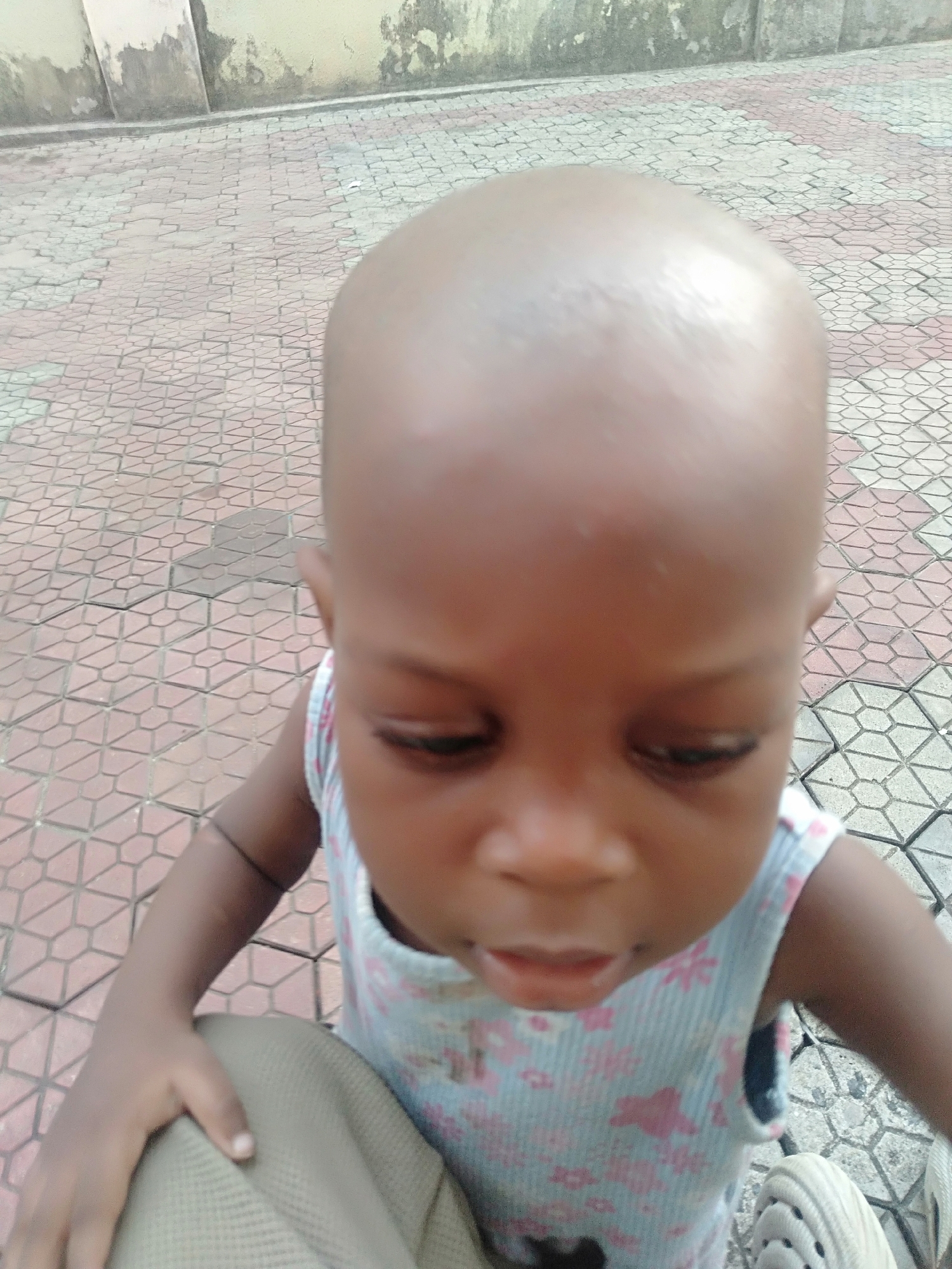 A bald, young child stands on a patterned tile surface. The child wears a light blue and pink floral patterned sleeveless shirt. The background includes a faded wall. Based on the request, this is in Shangisha, Nigeria.