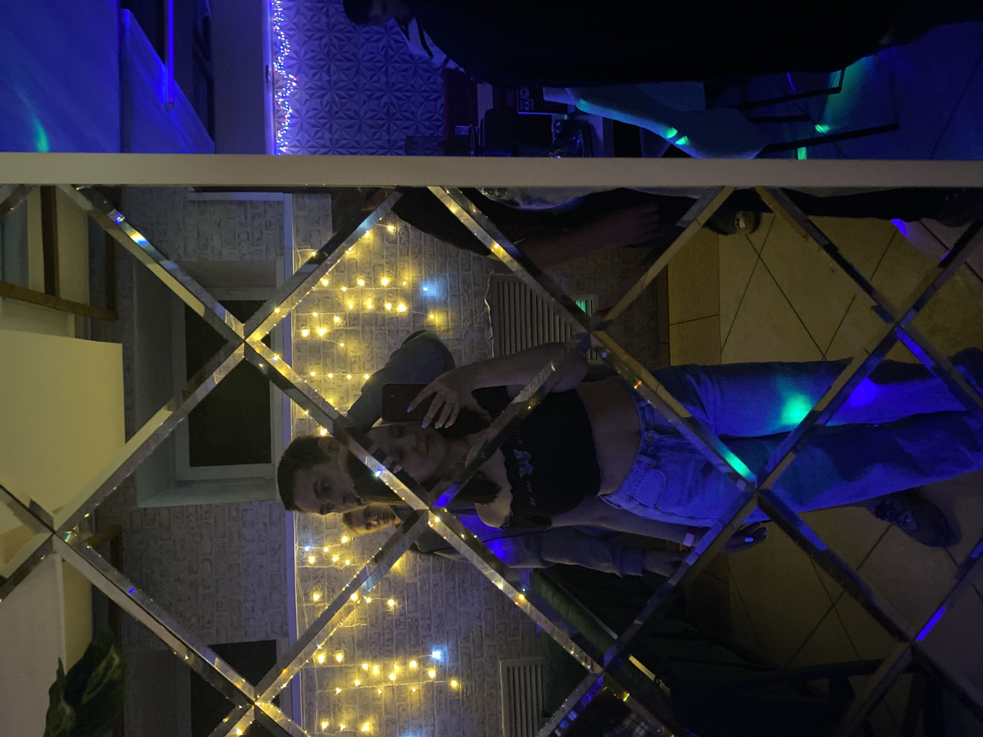 A mirror framed in a diamond pattern reflects a young couple taking a selfie in a room illuminated by string lights. The female is holding a smartphone. The male has his arm around the female. The room has a wall with a textured design. The image also shows the ceiling above the couple and floor beneath them. This appears to be a photograph taken in the city of Chaikovsky, Russia.