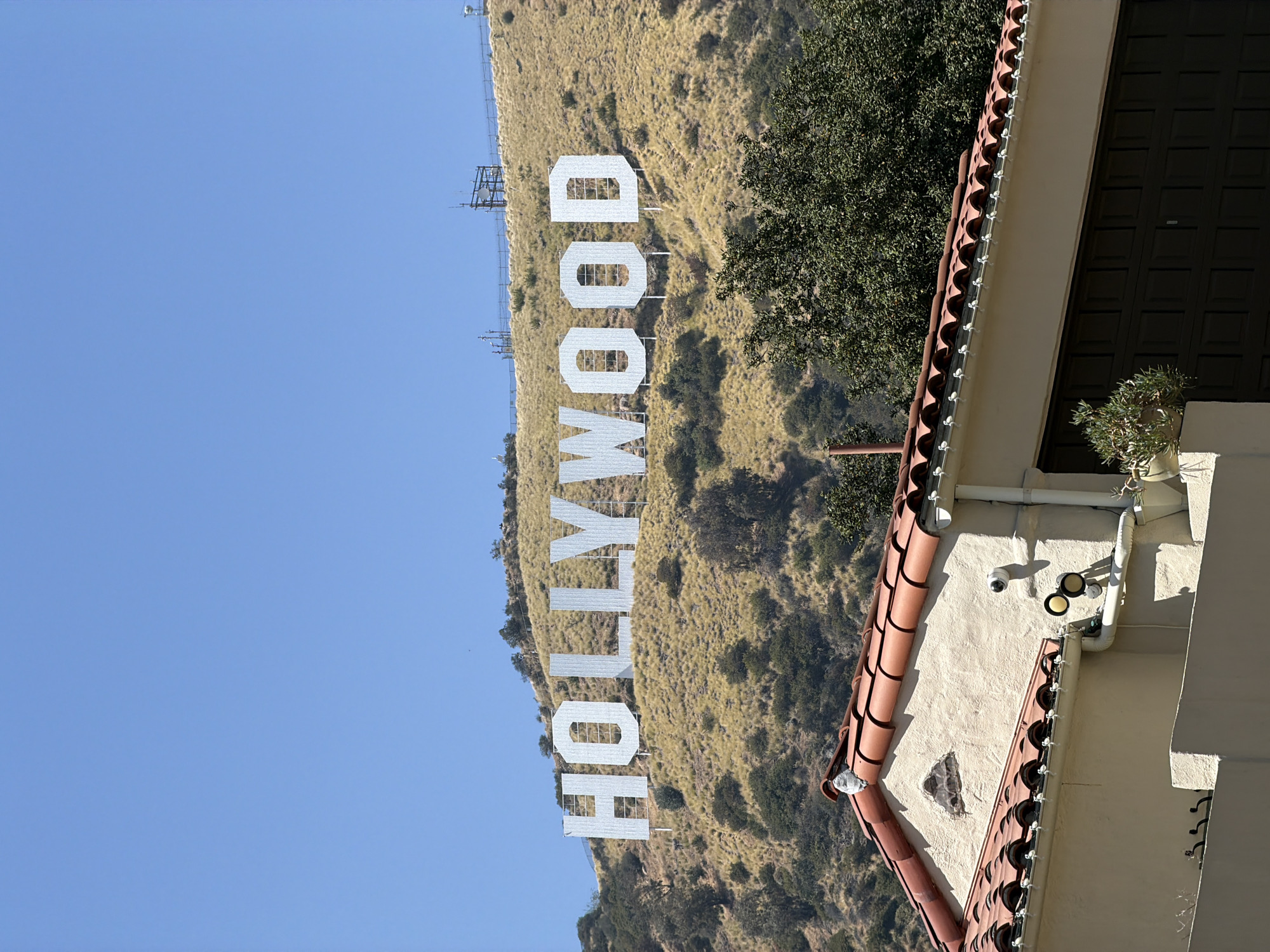 The image depicts the Hollywood Sign in Los Angeles, California. The sign is situated on a hillside covered in dry grasses and scattered trees. The sky is clear and blue. In the foreground, the edge of a building with a red-tiled roof and beige exterior is visible. A security camera is mounted on the building's exterior wall. A potted plant sits on a ledge near the wall.