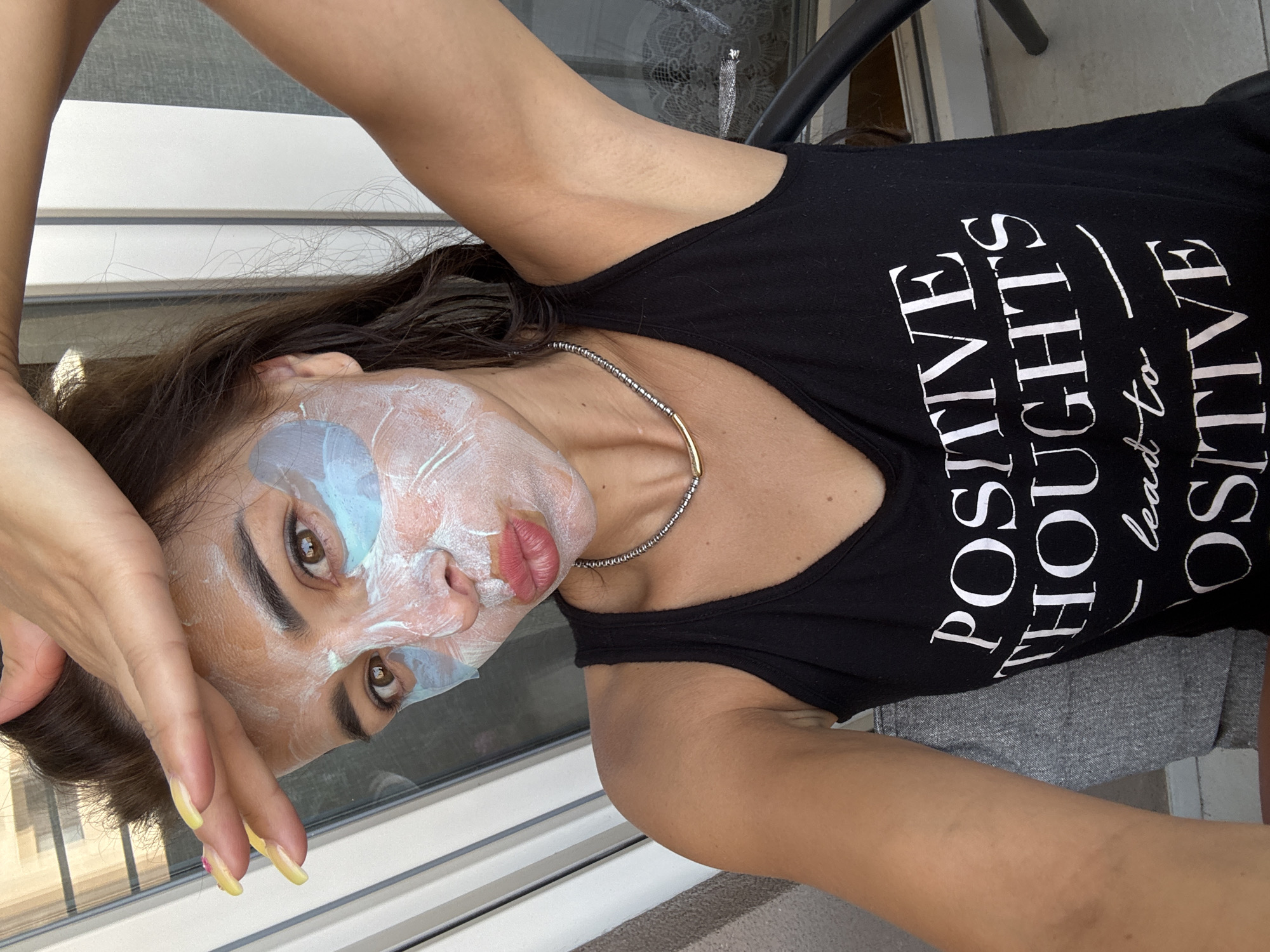 A woman is shown in a close-up selfie, applying a face mask. She has dark hair, brown eyes, and is wearing a black tank top with white lettering that reads "POSITIVE THOUGHTS lead to POSITIVE". She has blue under-eye patches and a white mask on her face. Her left hand is raised to her forehead, and her right arm is raised. The background appears to be outdoors, with vertical blinds visible. The location is Unknown, Unknown.