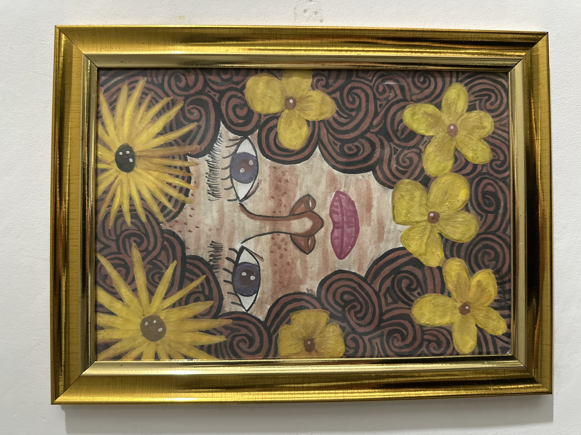 A framed painting hangs on a white wall. The painting depicts a stylized face surrounded by curly brown hair, yellow flowers, and swirls. The frame is gold-colored. The image does not indicate the location of Davao City, Philippines.