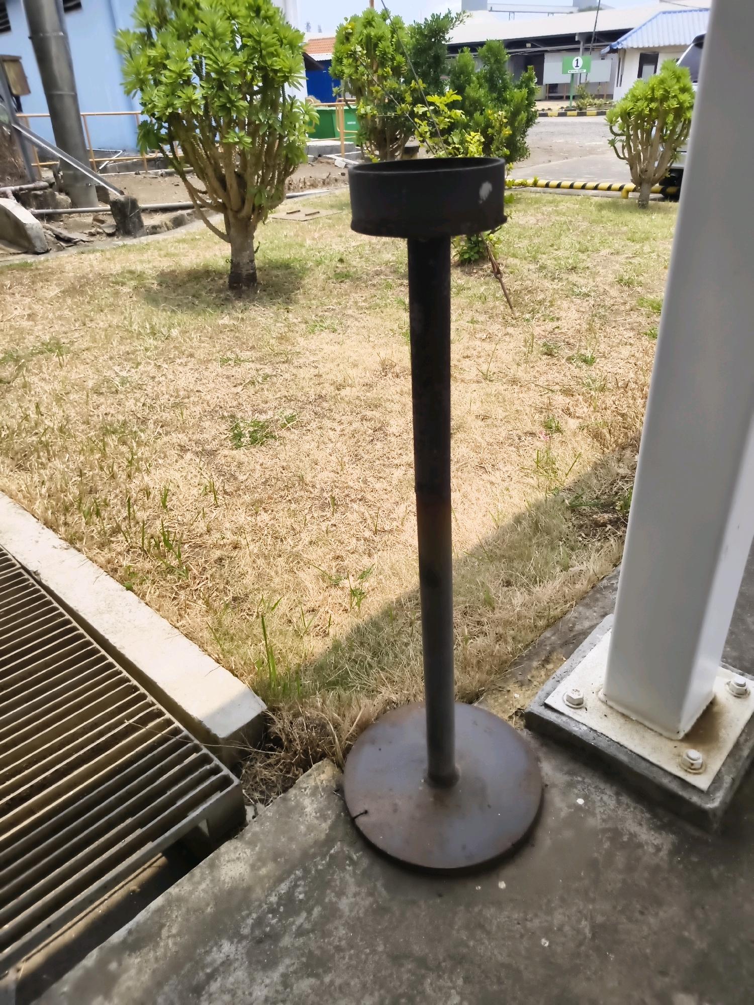 The image shows an outdoor scene with a black ashtray stand in the foreground. The stand has a circular base and a tall, slender pole with an ashtray receptacle at the top. To the left of the ashtray, a metal grate covers a drainage channel. The ground is covered in dry, yellowish grass with some sparse green shoots. In the background, there are several small trees and a building with a blue roof. The scene appears to be in an industrial or commercial area. The location is specified as Salira, Indonesia, though there are no specific landmarks visible to confirm this.