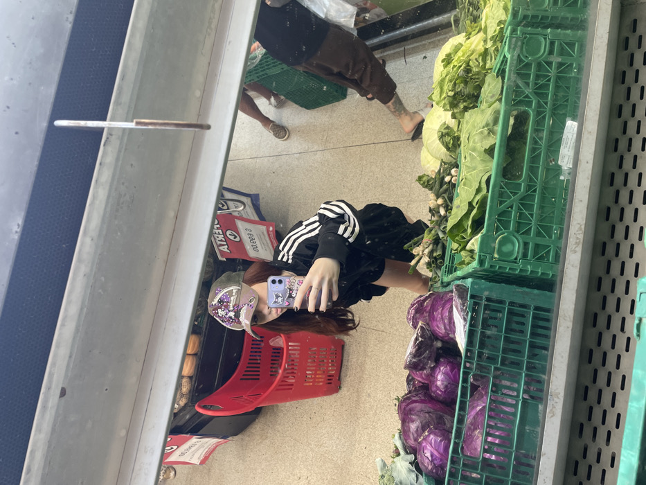 A mirror reflects a scene inside a grocery store. A person with long brown hair, wearing an Adidas tracksuit and a hat, holds a phone to take a selfie. There's a red shopping basket beside them. Crates of green leafy vegetables and purple cabbages are visible. Reflected above is another person wearing shorts and no shoes.