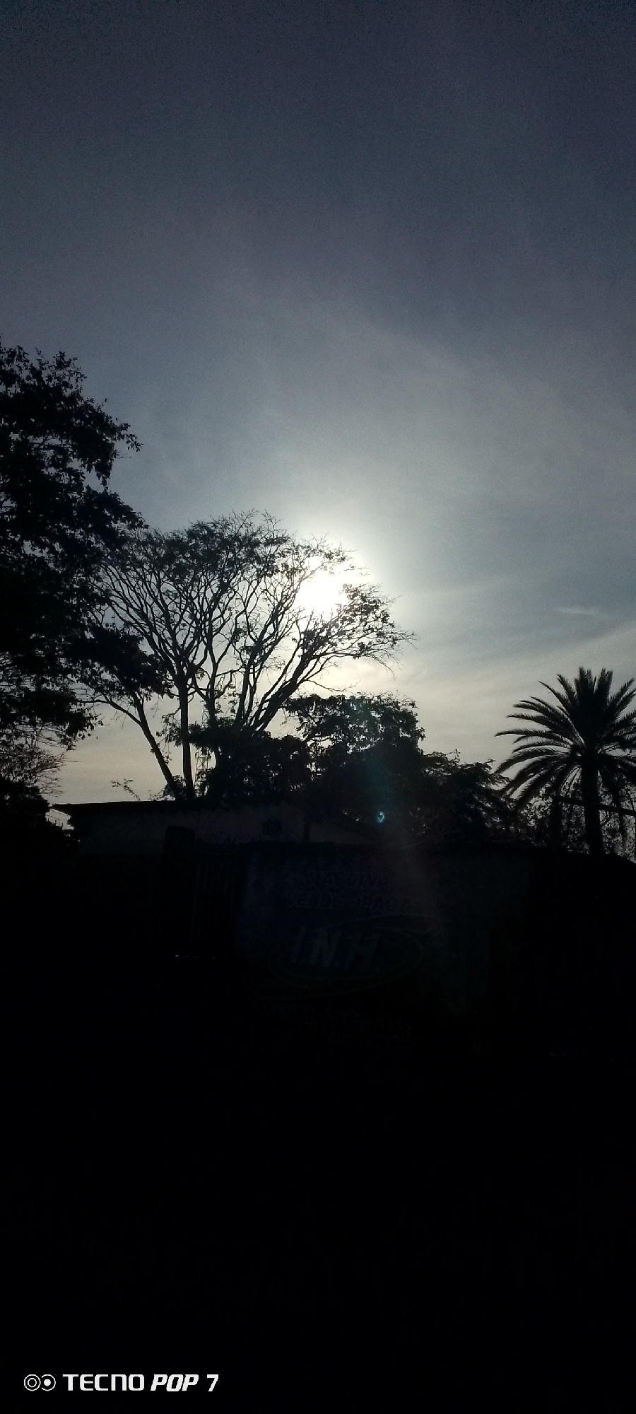 The image shows a silhouetted scene, likely in Altagracia, Venezuela, given the prompt. The sky is overcast with the sun obscured by clouds, creating a hazy effect. The foreground is dark, concealing details. Visible above the darkness are trees, including a palm tree on the right. A building is faintly visible behind the trees. The bottom of the image displays the text "TECNO POP 7," indicating the type of camera used to capture the photo.