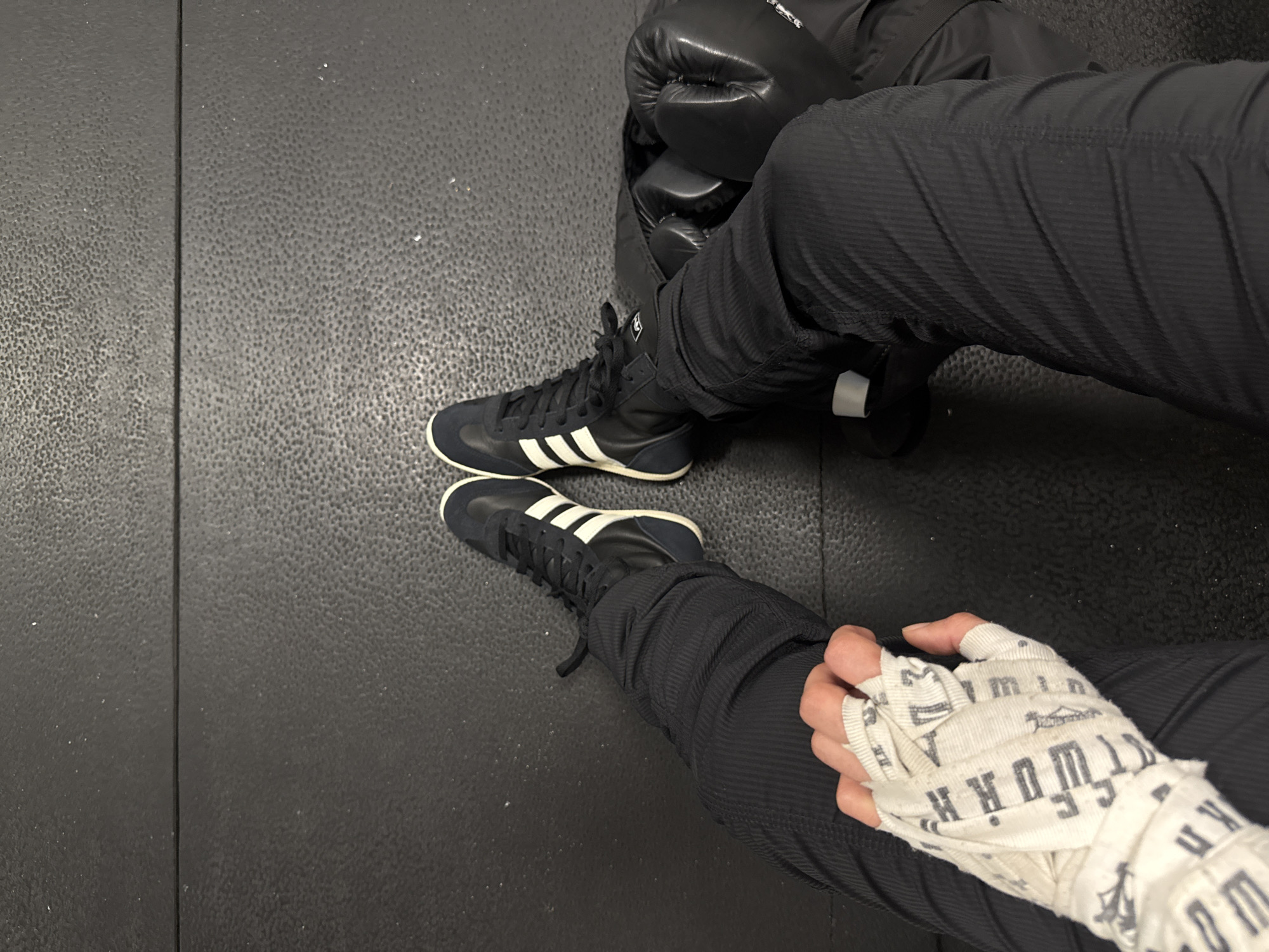 The image depicts a person seated on a black, textured floor, possibly a gym mat. The person is wearing black athletic shoes with three white stripes, indicative of the Adidas brand, and black pants. Their right hand is wrapped in white boxing hand wraps, suggesting preparation for a boxing or martial arts session. Next to the person's left leg is a pair of black boxing gloves and a dark-colored athletic jacket. The location is indeterminate, but there is no evidence to indicate this is in London, United Kingdom. There are no specific features or details pointing to London.