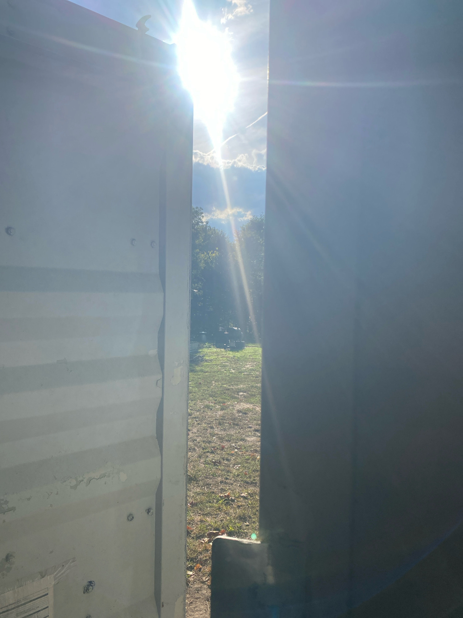 An outdoor scene is captured from the perspective of looking through a gap between two large, light-colored, corrugated metal panels. The panel on the left is textured with horizontal ridges and shows some peeling paint and visible fasteners. The panel on the right is a solid, darker gray surface.
Through the opening, a sun-drenched landscape is visible. The sun is very bright, causing lens flare and obscuring details in the sky, which contains scattered clouds and a contrail. Rays of sunlight emanate from the sun, creating a strong backlight.
Beyond the panels, a grassy area with scattered dry leaves is present. In the middle ground, a cluster of trees with green foliage can be seen. There are some indistinct objects in the yard, possibly outdoor equipment or structures. The overall impression is a bright, sunny day in a semi-rural or outdoor setting. The location is identified as Old Fort, United States.