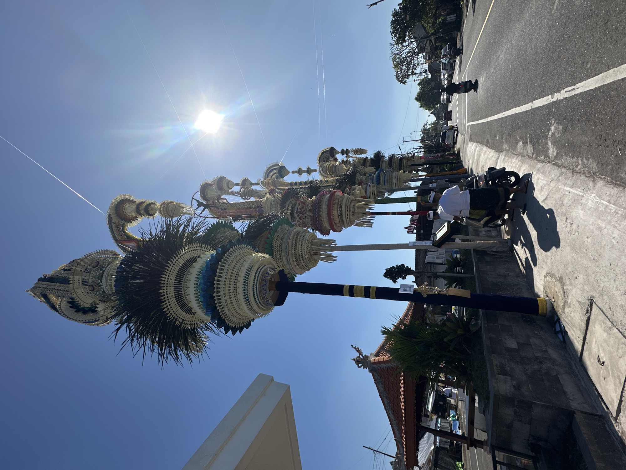 The image depicts a street scene in Mengwi, Indonesia, under a bright, clear blue sky with the sun prominently featured and creating lens flare. On the left side of the road, a row of tall, ornate decorations, likely religious or ceremonial in nature, are erected on poles. These decorations are intricately woven and layered, featuring a variety of textures and colors, including white, gold, blue, and dark green. Some of these structures resemble palm fronds and woven straw elements. In the foreground, a person wearing a white shirt and a dark sarong is standing next to a parked motorcycle, facing the road. To the left, a section of a white building with a sharp angle is visible, and further back, a traditional Balinese-style building with a red tile roof and intricate carvings is present, surrounded by greenery. A stone wall runs along the edge of the sidewalk. The road itself is asphalt, with a white dividing line. Several vehicles, including motorcycles and cars, are visible on the road, with two motorcycles actively being ridden. The environment suggests a warm, sunny day in a tropical urban or semi-urban setting, possibly during a festival or cultural event, given the elaborate decorations.