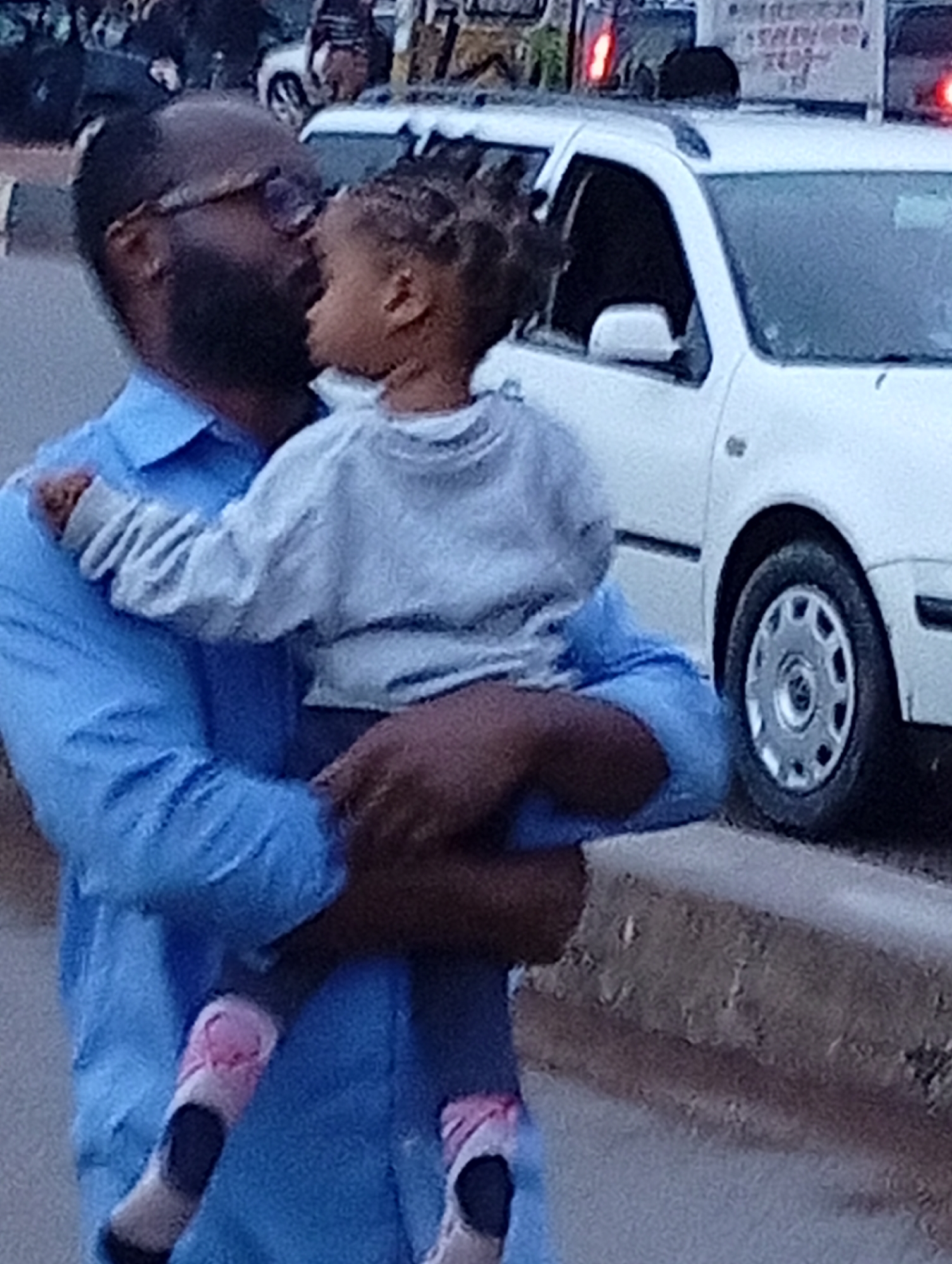 In Jos, Nigeria, a man in a blue collared shirt is holding a young child in his arms. The child is wearing a light blue long-sleeved shirt and pink and white patterned socks. The man has a beard and is wearing glasses. They are standing in front of a white SUV with its doors slightly ajar. The background shows a street with a sidewalk and hints of other vehicles and structures. The lighting suggests it is daytime. The man appears to be kissing or nuzzling the child.