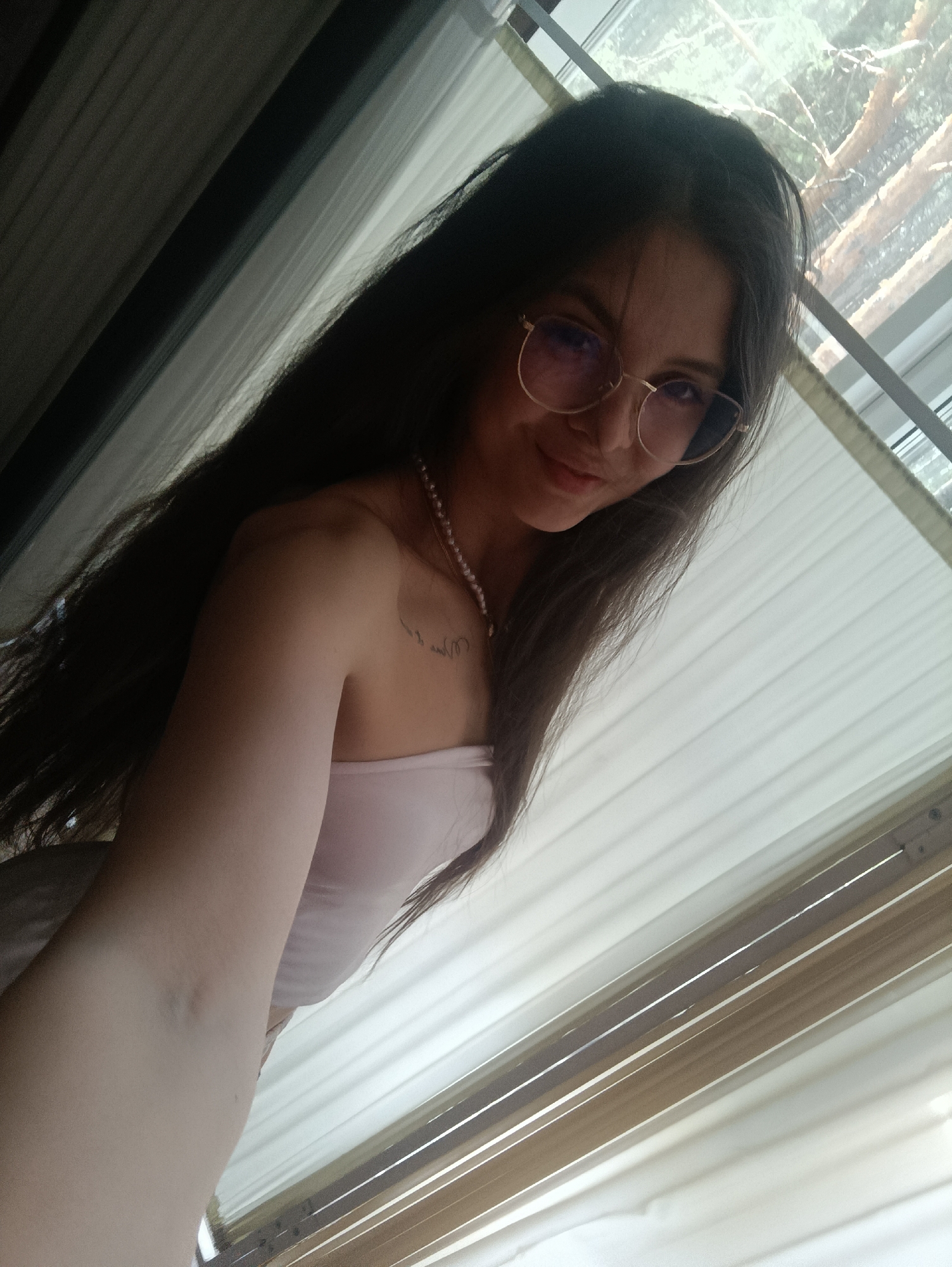 A woman with long, dark hair is seen in a selfie-style image, wearing round gold-rimmed glasses, a pearl necklace, and a light pink strapless top. She is positioned in front of a window with white blinds, which are partially open, revealing green foliage or trees outside. There is a tattoo on her shoulder, and the arm holding the camera is visible in the foreground. The location is Unknown, Unknown.