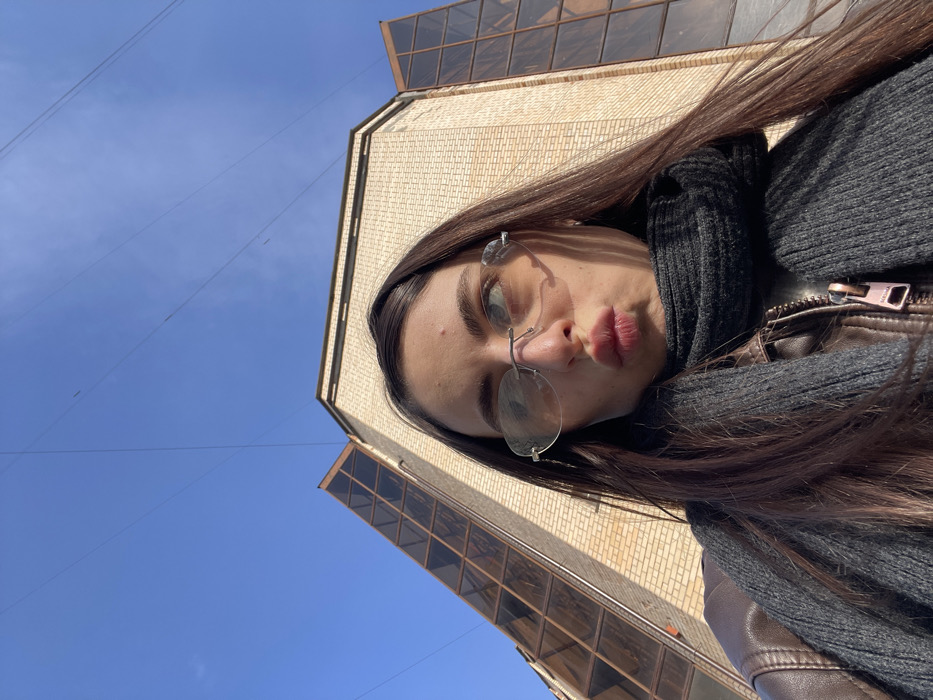 A female subject, positioned for a low-angle selfie, is captured outdoors in bright daylight. She has long, dark hair parted centrally and is wearing a dark brown leather jacket and a dark grey ribbed knit scarf. Her face is turned slightly to the right with lips pursed. She wears rimless, amber-tinted sunglasses featuring decorative silver accents on the temples. A small silver earring is visible on her right ear. The background features a distinctive multi-story building constructed with light beige brickwork. The building exhibits angular, faceted architecture, with prominent dark, segmented, sloped glass panels on the left side and a flatter dark glass section on the right. Above the building, a clear blue sky is visible, traversed by several thin, dark electrical wires. The building is identified as the Trade House "Pravda" (Торговый дом "Правда"), located at ulitsa Pravdy, 24, building 3, in Moscow, Russia.