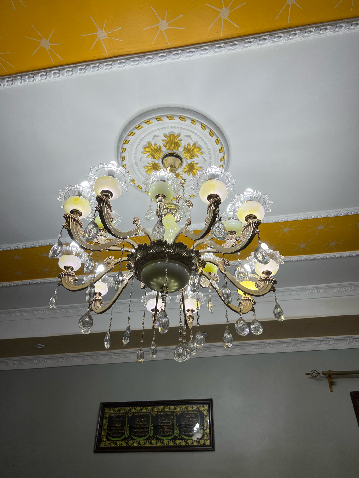 A chandelier hangs from a white ceiling with ornate gold and white molding. The ceiling is painted yellow in a wide band below the molding, with a pattern of white sunbursts. The chandelier has multiple arms holding frosted glass globes and is adorned with numerous clear crystal teardrop pendants. Below the chandelier, on a gray wall, hangs a framed piece of Islamic calligraphy. The scene appears to be an indoor residential or public space. The city of Dar es Salaam, Tanzania, is not visually identifiable from the image.