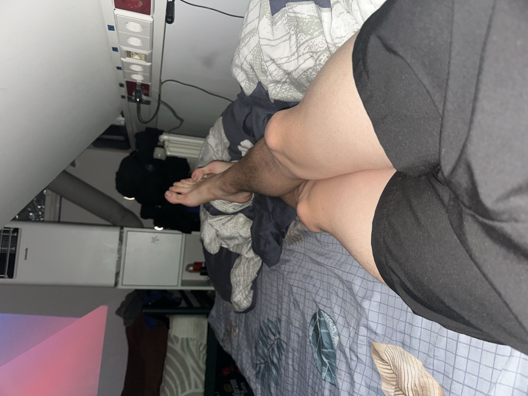 The image shows a person's legs and feet resting on a bed covered with a blue checkered duvet featuring a leaf pattern. The person is wearing black shorts. In the background, there is a white portable air conditioner, electrical outlets, and a radiator. The scene is likely in a bedroom, possibly in a city like Mainz, Germany, given the presence of European-style electrical outlets. The lighting is dim, suggesting it might be nighttime or a room with limited light sources.