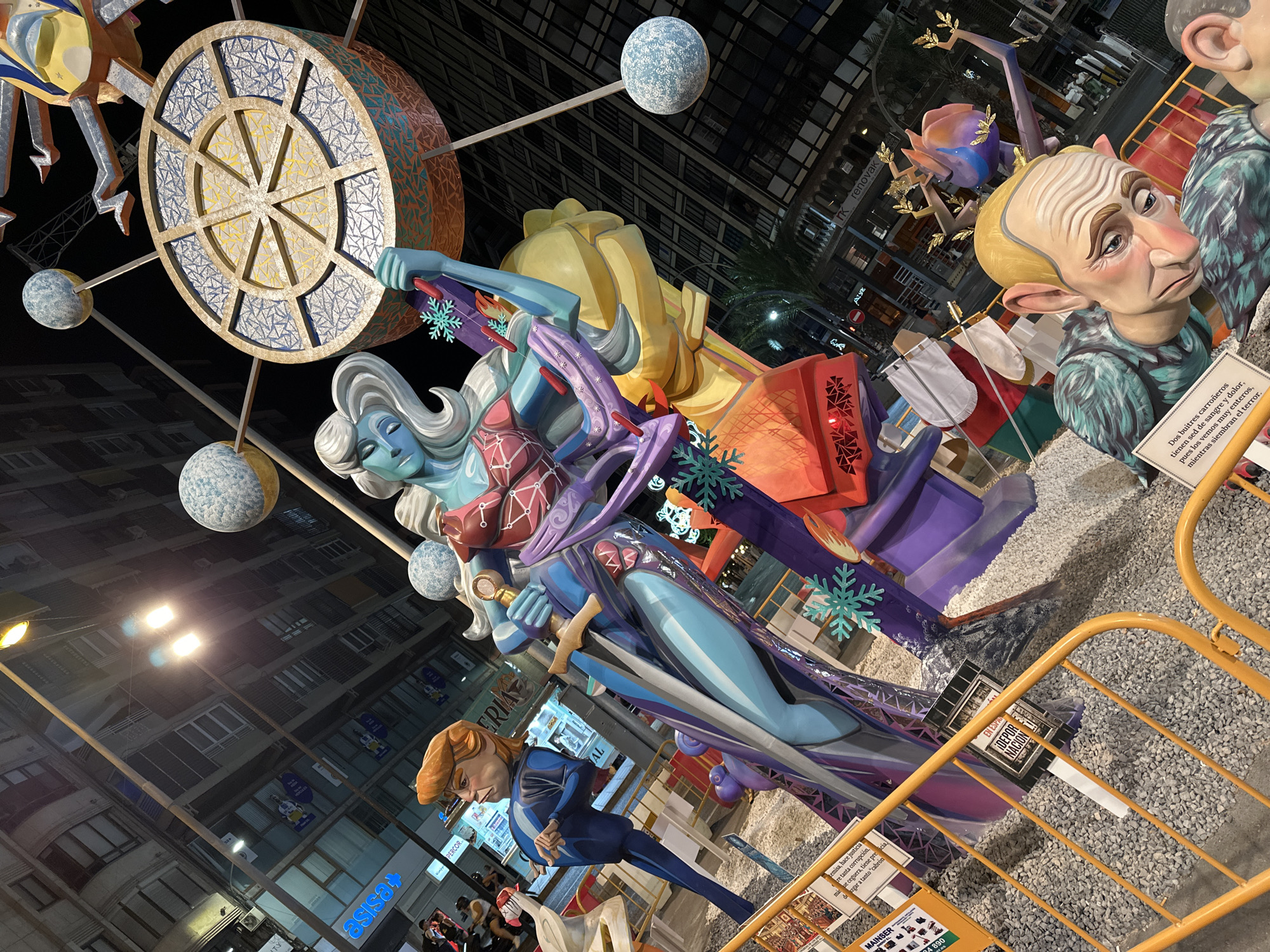 The image depicts a large, elaborate street installation, likely part of a festival, in Alicante, Spain. The artwork features several oversized, cartoonish figures. In the foreground, a large, grotesquely rendered figure with a bald head, wrinkled skin, and a pensive expression is visible, depicted with a turquoise feathered garment. Behind this figure, a tall, slender figure with light blue skin and white hair, wearing a stylized red and blue outfit, holds a sword. To the left of this figure is another character with orange hair and a blue suit. The installation includes various decorative elements such as spheres resembling planets, snowflakes, and a large circular structure with intricate patterns. The background reveals a city street at night with buildings, lights, and shops, including a sign that reads "asisa". Yellow safety barriers are placed in front of the artwork, with informational signs attached. The ground is covered with small gray stones.