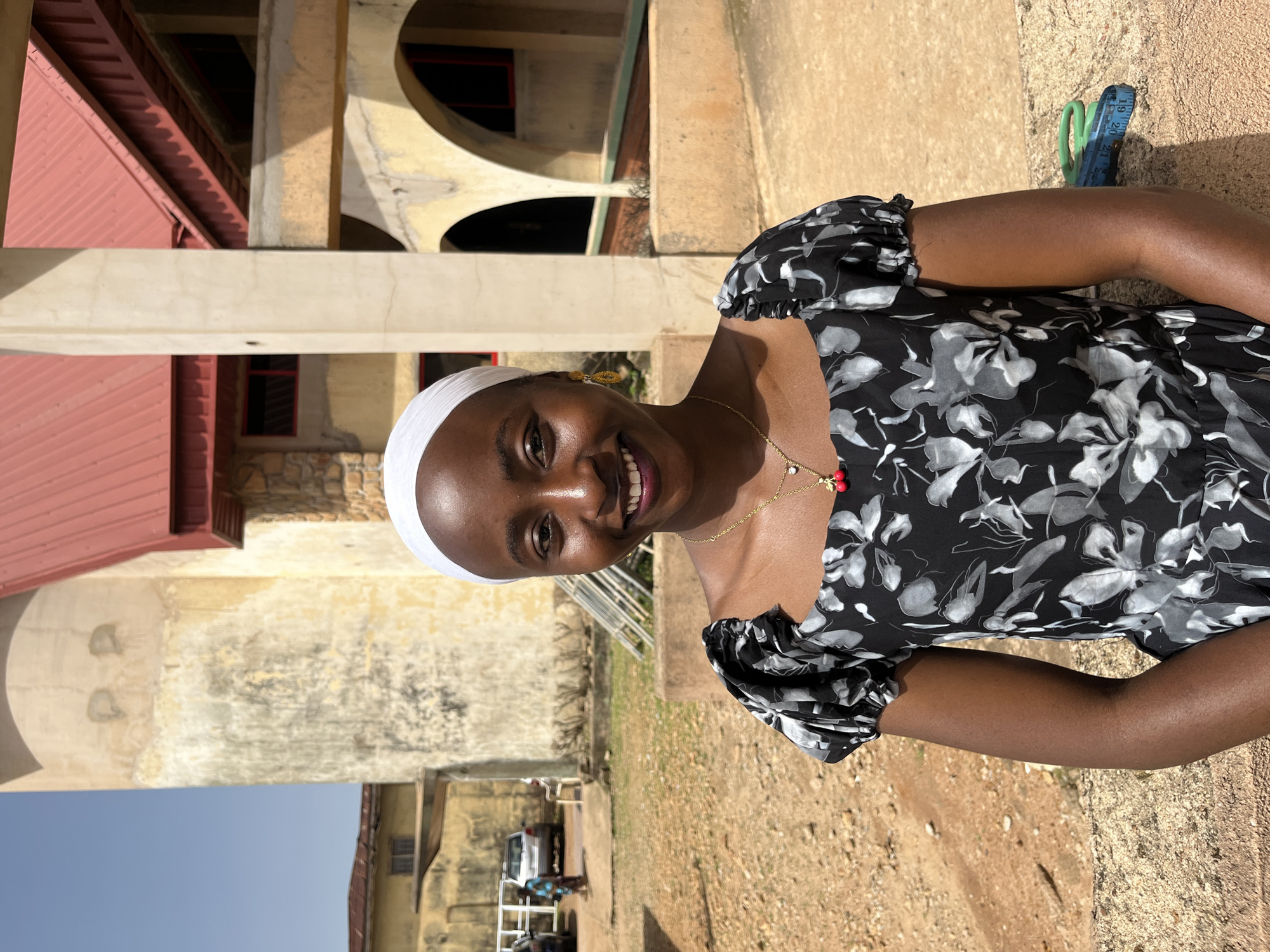 A medium shot captures a young Black woman standing outdoors in Bukuru, Nigeria. She is smiling directly at the camera, her head tilted slightly. She is wearing a white headscarf and a black dress with a floral pattern. A gold necklace with a red pendant hangs around her neck. The background shows a building with a red roof and beige walls, some arches, and a tall, weathered concrete tower. The ground is covered in dirt and gravel. Several vehicles and people are visible in the distance, suggesting an urban or semi-urban environment. A measuring tape and scissors are on the ground to the right of the woman.