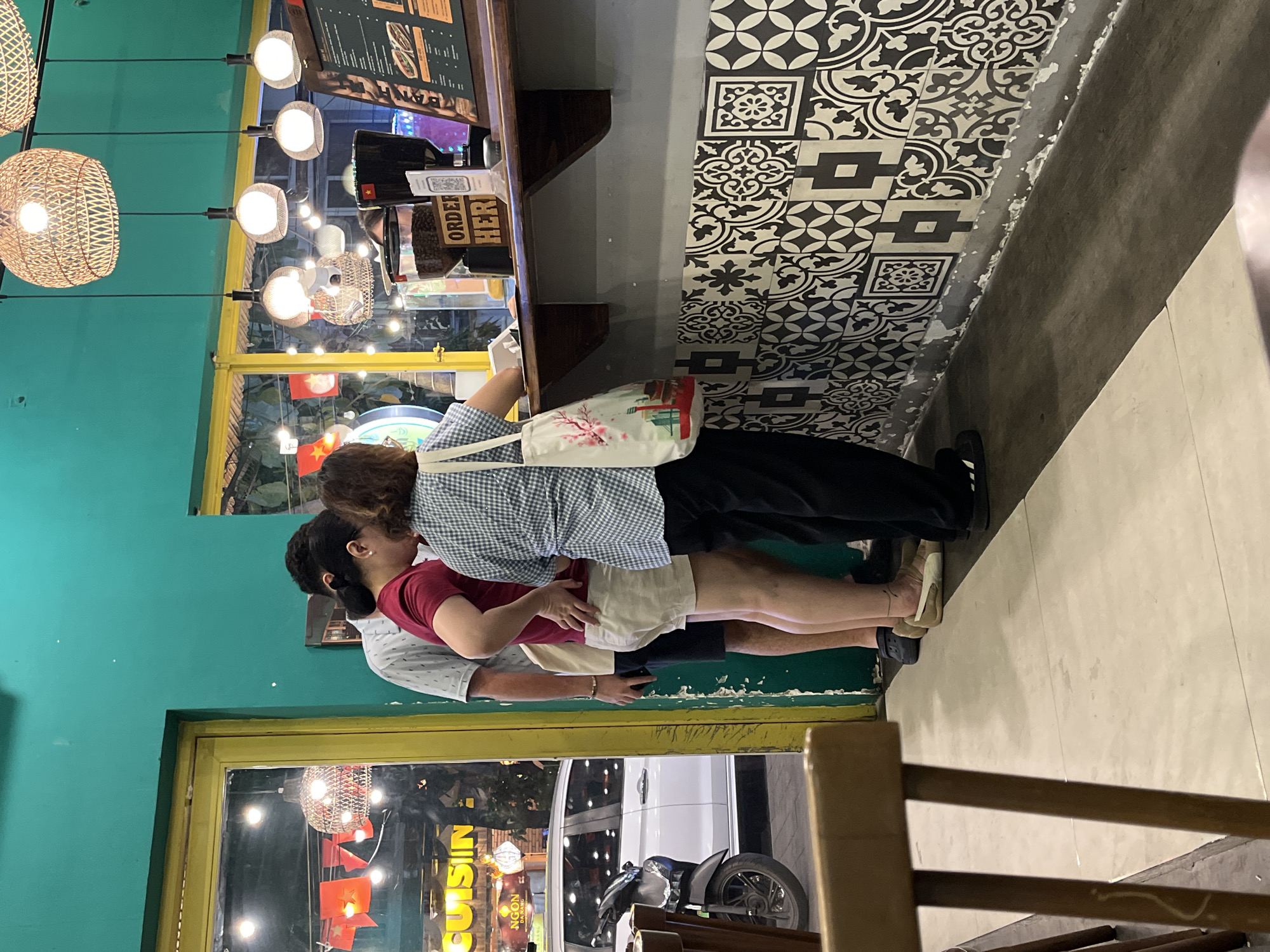 The image captures a scene inside a restaurant in Đà Nẵng, Vietnam. Three people stand in line at the order counter. The person closest to the counter is wearing a light blue checkered shirt, black pants, and a white bag. The second person in line is wearing a red shirt and light-colored shorts. The third person is wearing a white shirt. The order counter is next to a black and white patterned wall. A window is visible behind the people, displaying the exterior of the restaurant and a view of the street, which includes a van and a parked motorbike. The restaurant is lit by hanging wicker lamps. Signs with "CUISINE" are visible behind the window.