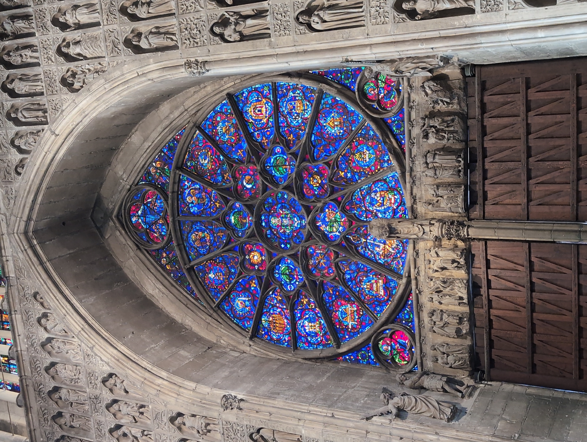 The image shows a large, intricately designed rose window above a pair of massive wooden doors, part of a grand stone building, likely a cathedral or church. The rose window is filled with vibrant stained glass, predominantly in shades of blue, red, and yellow, forming geometric patterns and figural scenes. Surrounding the rose window and on the facade of the building are numerous stone sculptures of figures, arranged in niches and rows. These figures appear to be religious in nature, possibly saints or biblical characters, depicted in various poses and with distinct clothing. The wooden doors are imposing and appear to be ancient, with a rustic, planked design. The overall impression is one of significant architectural and artistic detail, indicative of a historical and possibly religious site. The location is identified as Urvillers, France, though without further context or specific landmark identification within Urvillers, the exact building cannot be pinpointed.