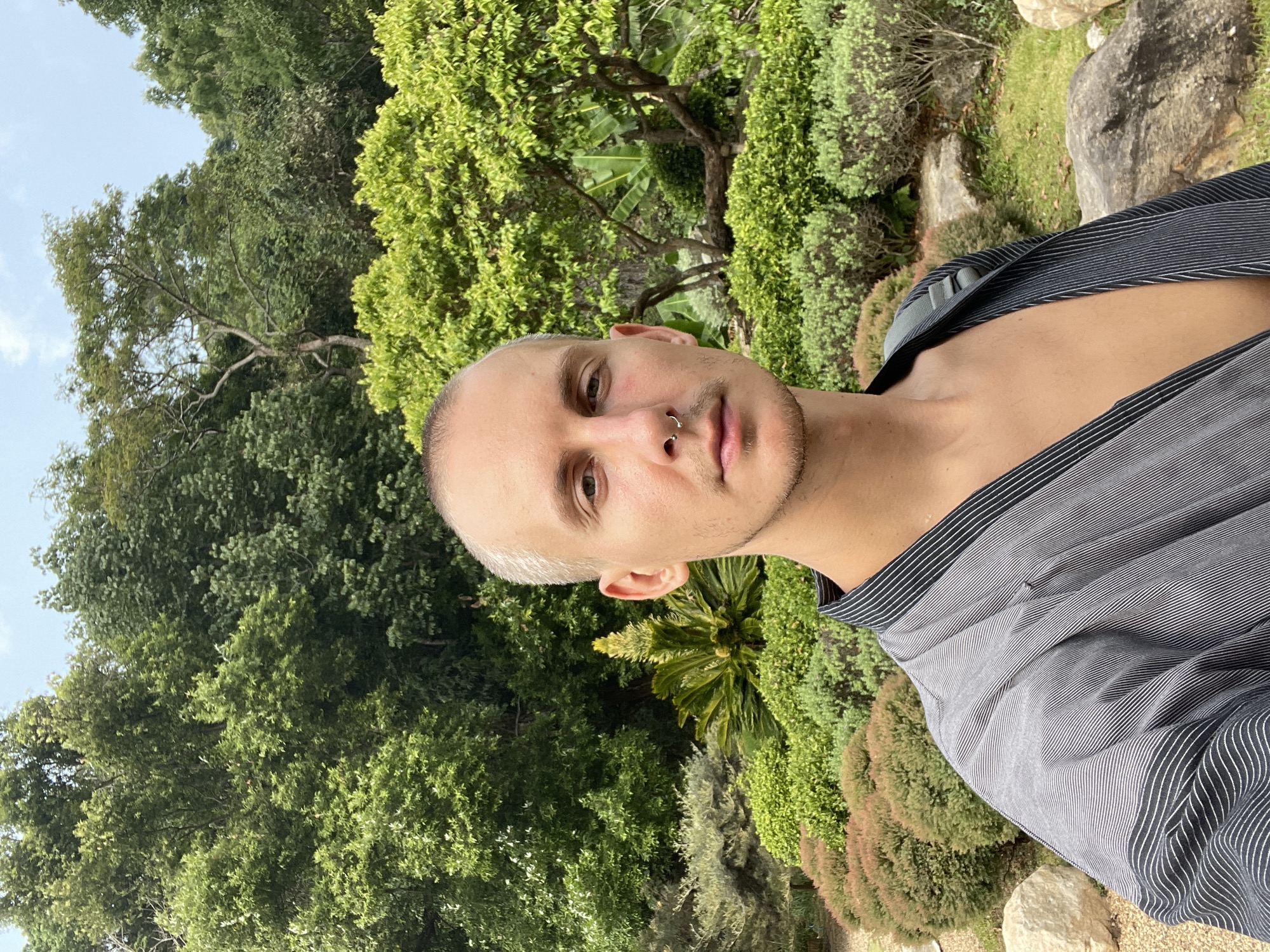 This media file features a close-up, rotated image of a young adult male in an outdoor, lush green environment. The image is rotated 90 degrees clockwise, showing the man's head on the left side of the frame and the sky in the upper left corner.
The subject is a man with very short, light-colored hair and light eyes. He has a subtle septum piercing (a silver hoop) and light stubble on his face. He wears an open-collared, dark grey or black shirt with thin white vertical stripes, which is pulled down on his left shoulder, exposing part of his chest and shoulder. He looks directly at the camera with a neutral and direct expression, suggesting he is posing for a selfie or a portrait.
The background is dominated by dense, vibrant green foliage, including various trees and shrubs, indicative of a garden or natural park. Some larger, broad-leafed plants are visible, consistent with a tropical setting. In the lower right of the frame, large rocks and patches of grass can be seen, marking the ground. The upper left corner reveals a bright blue sky with a few faint white clouds, suggesting clear and sunny weather during daytime. The strong natural light illuminates the scene evenly. No visible text is present in the image. The setting aligns with the provided location context of Ban Nong Hoi, Thailand.