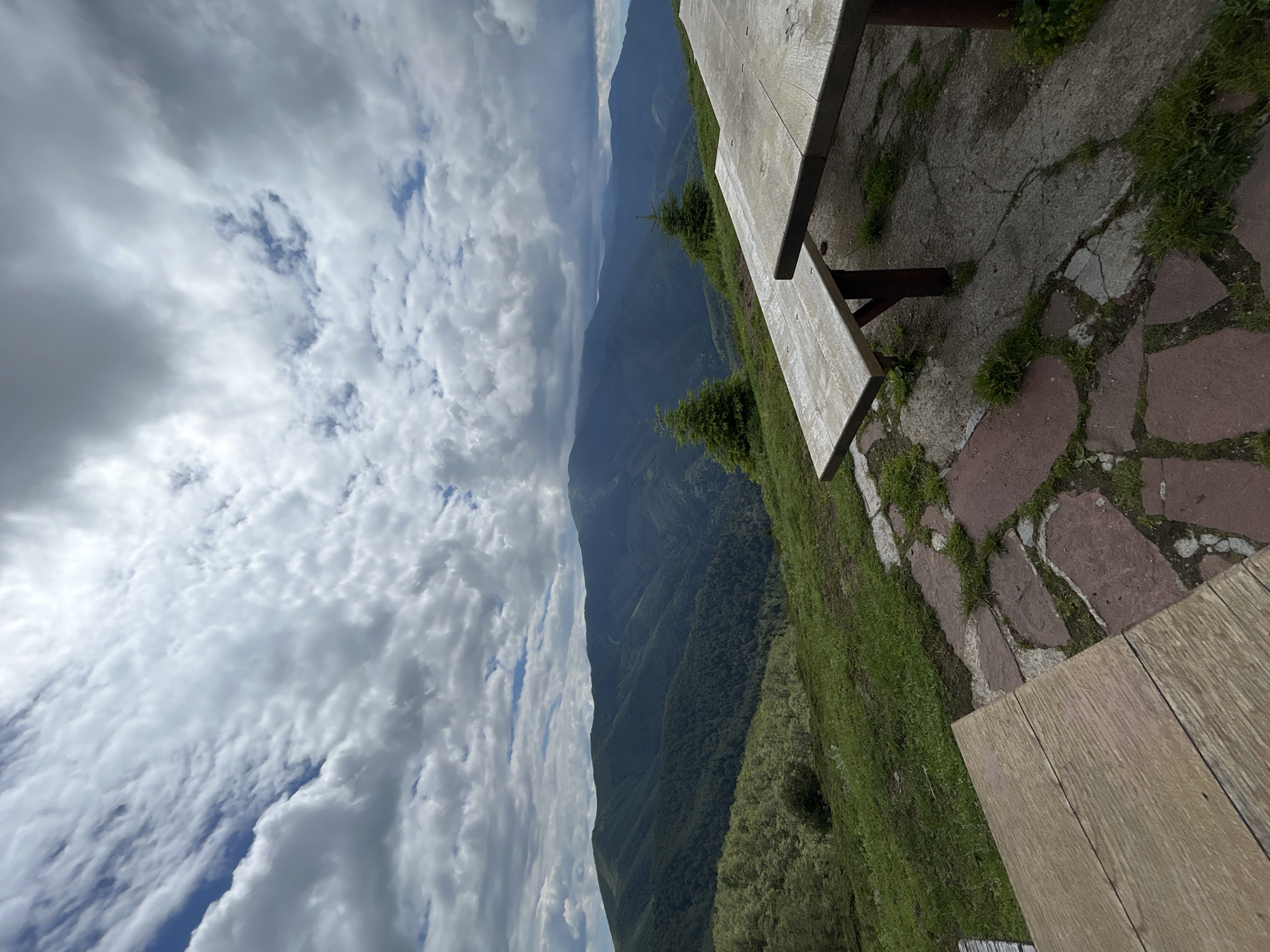 The image depicts a scenic view of mountains and a cloudy sky from a picnic area. In the foreground, there are two rustic wooden picnic tables with benches. One table is positioned closer to the viewer and takes up the lower left portion of the frame, while the other table and bench extend into the right side of the image. The picnic area is situated on a slight incline covered with green grass and interspersed with a stone pathway made of irregular, reddish-brown and grey stones. Beyond the picnic area, the landscape rolls into a series of lush green, forested mountains stretching into the distance under a dramatic sky filled with white and grey clouds. Two small evergreen trees are visible on the grassy slope between the picnic tables and the mountains. This location is identified as being in the city of Rozino, Bulgaria.