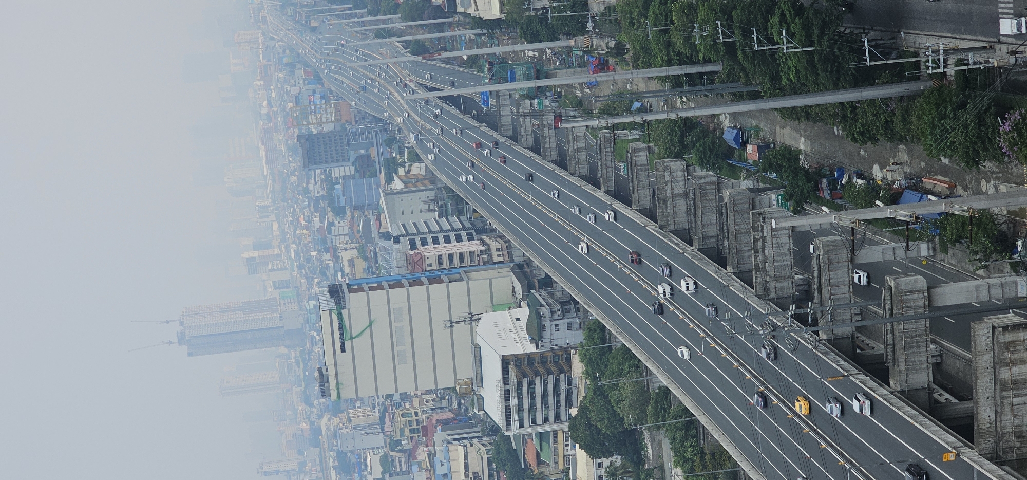 An aerial, eye-level view captures a multi-lane elevated highway stretching into a hazy cityscape of Makati, Philippines. The highway, bordered by white lane markers, carries a moderate flow of vehicles including cars, vans, and a yellow taxi. Construction elements, such as concrete supports and scaffolding, are visible along the side of the highway. Adjacent to the highway, residential and commercial buildings of varying heights and architectural styles are present. The distant skyline is obscured by atmospheric haze, with tall buildings and construction cranes visible through the haze. Trees and vegetation are interspersed throughout the scene, particularly along the edges of the highway and in pockets between buildings. The overall atmosphere is overcast, suggesting a cloudy or smoggy day.