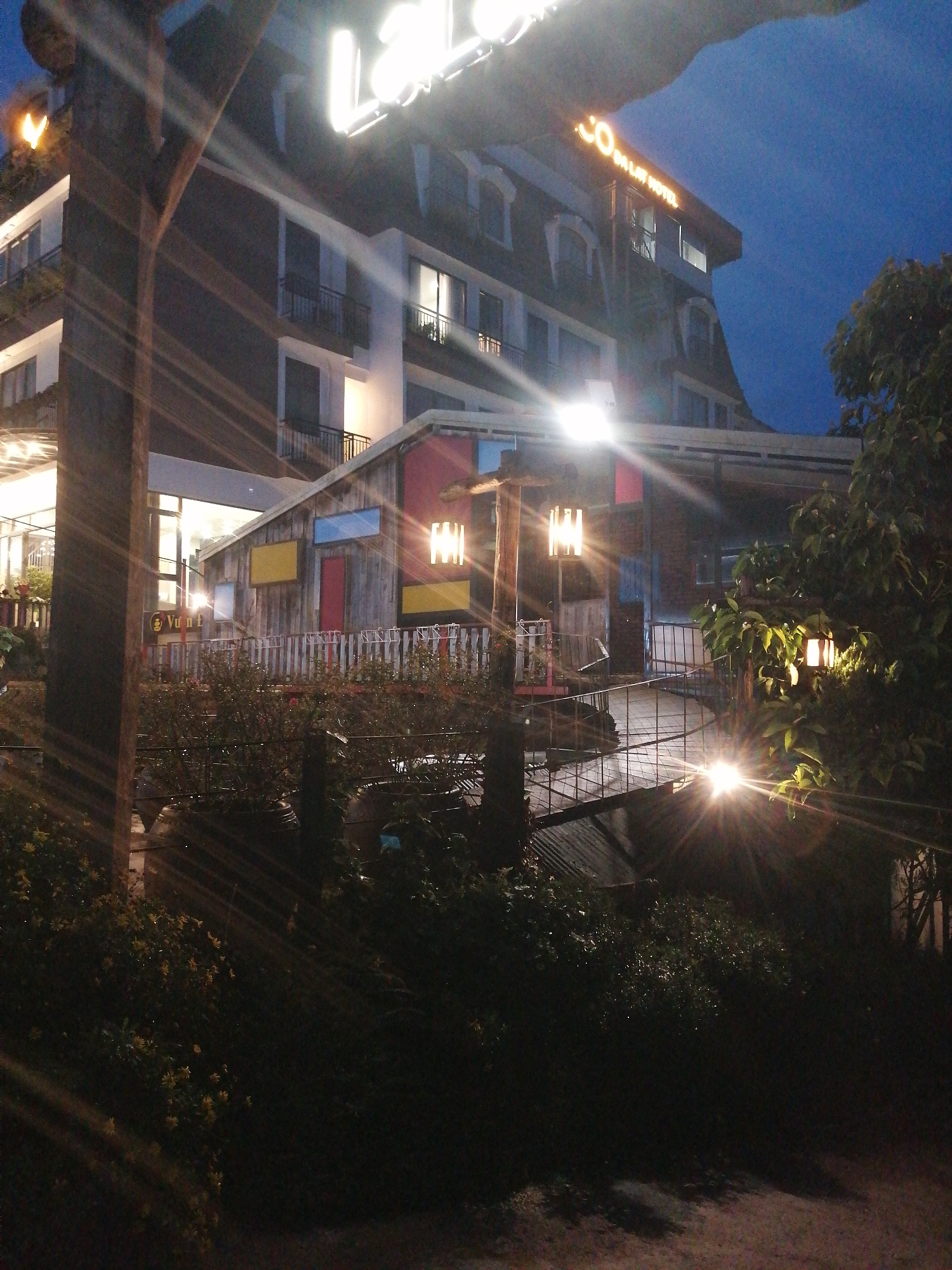 The image shows the exterior of the LaLa Land Hotel in Da Lat, Vietnam at night. A partially visible illuminated sign with "LaLa" is seen above the building. A sign on the top of the building reads "Co Do LaLat HOTEL". There is a wooden structure with brightly colored panels (red, yellow, blue) and a white picket fence in front of it. A small bridge with lanterns leads to this structure. Bushes and other greenery are located in the foreground and on the side of the image.